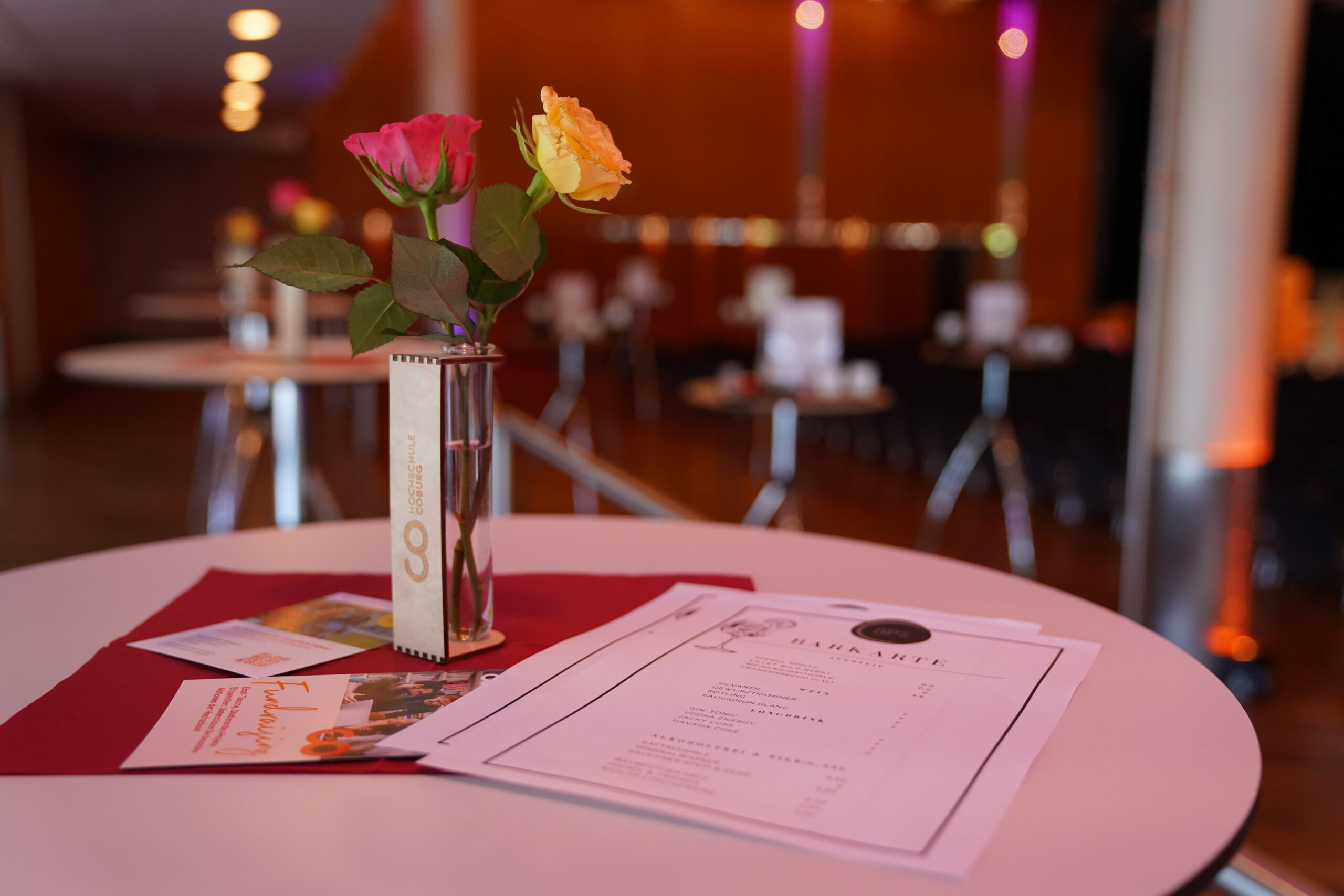 Eine Nahaufnahme eines runden Tisches mit Speisekarten, Broschüren und einer Vase mit zwei Rosen, die die feierliche Stimmung einer Absolventenverabschiedung 2025 in einem unvergesslichen, warm beleuchteten Veranstaltungsraum einfängt. at Hochschule Coburg
