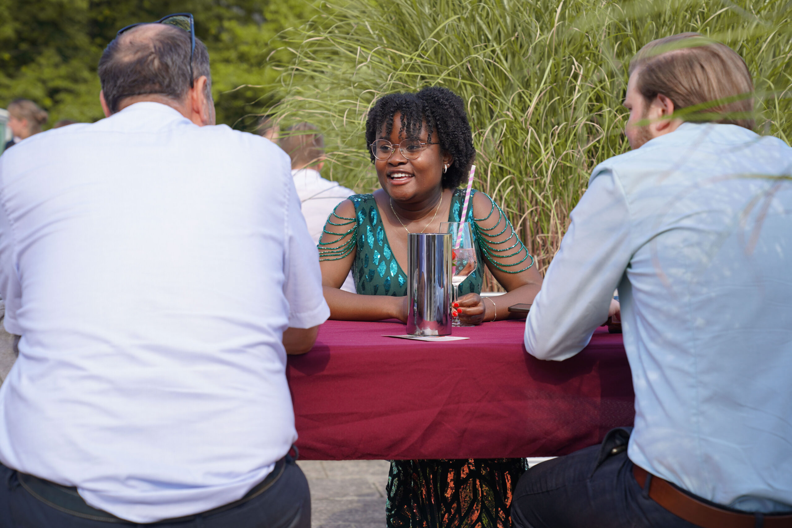 Eine Frau in einem grünen Kleid lächelt, während sie sich mit zwei Männern an einem Tisch im Freien mit einer kastanienbraunen Tischdecke unterhält. Im Hintergrund sind Gras und Grünpflanzen zu sehen. Die Frau hält ein Getränk in ihrer Hand. at Hochschule Coburg