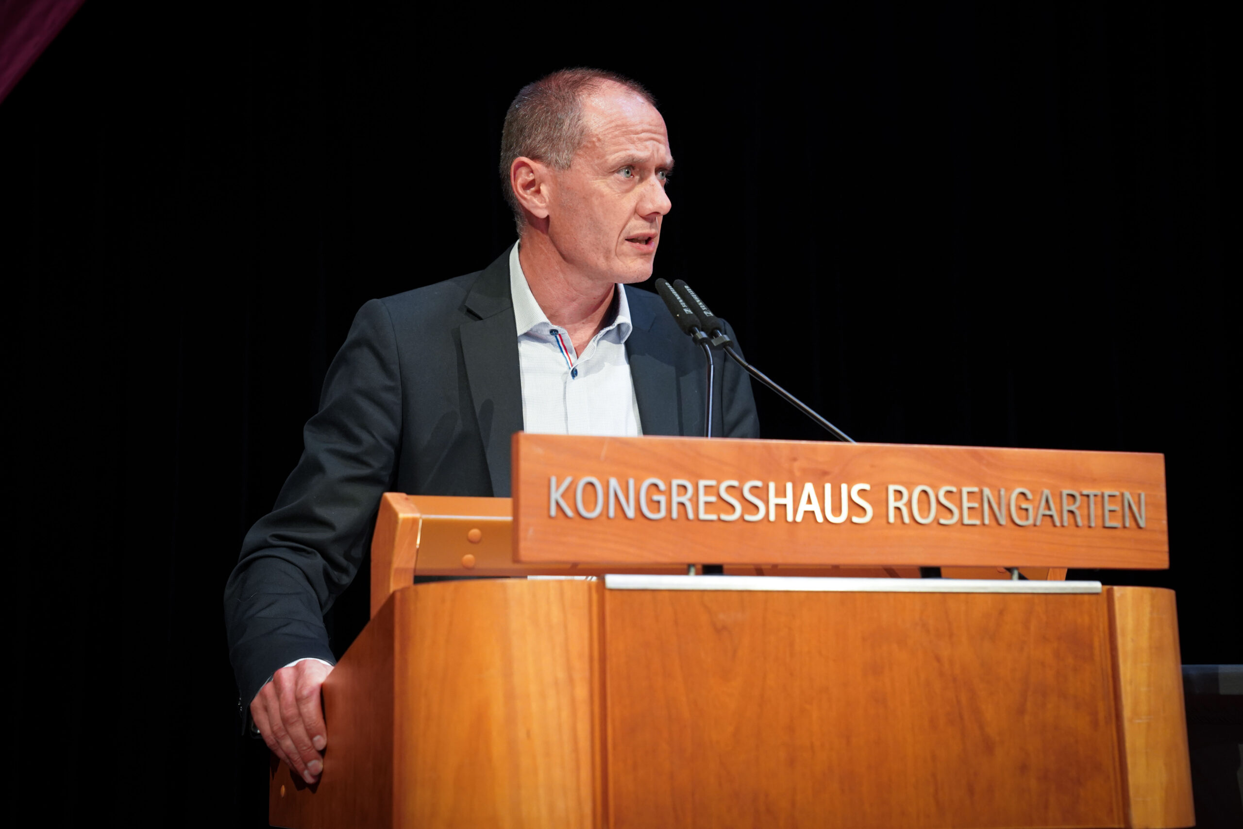 Ein Mann im Anzug spricht an einem Holzpodium mit der Aufschrift "KONGRESSHAUS ROSENGARTEN" vor einem dunklen Hintergrund. at Hochschule Coburg