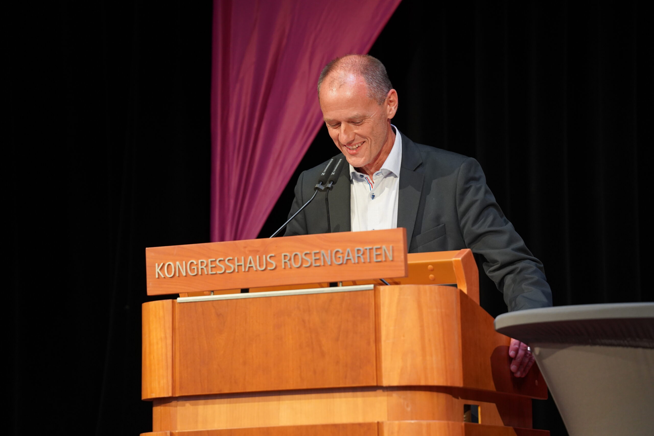 Ein Mann im Anzug steht an einem hölzernen Podium mit der Aufschrift "KONGRESSHAUS ROSENGARTEN", lächelt und schaut nach unten, während er während der feierlichen Absolventenverabschiedung 2025 in ein Mikrofon spricht. Im Hintergrund hängt ein drapierter rosa Stoff. at Hochschule Coburg