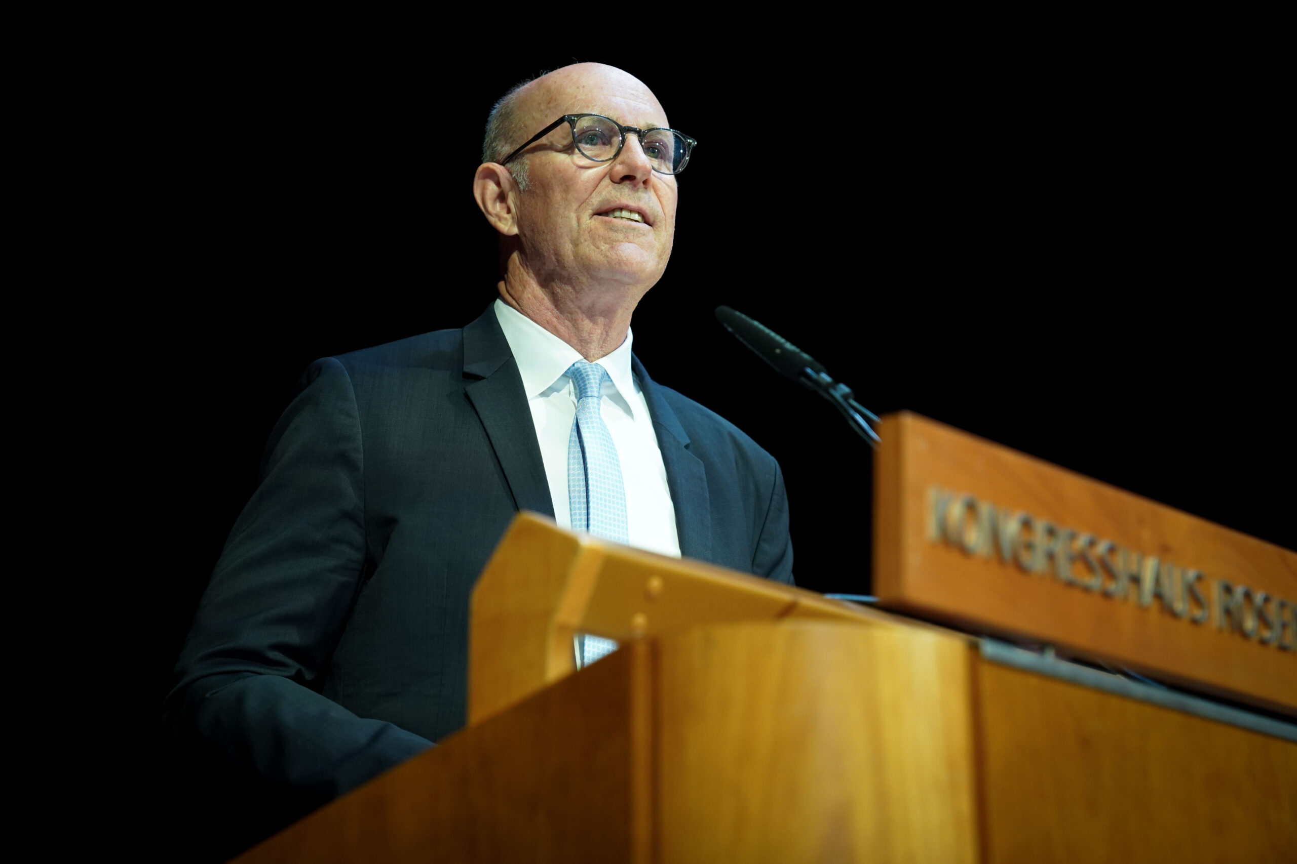 Ein Mann in Anzug und Krawatte steht an einem hölzernen Podium mit einem Mikrofon und spricht auf einer Veranstaltung. Der Hintergrund ist dunkel, und ein Teil des Wortes "KONGRESSHAUS" ist auf dem Podium zu sehen. at Hochschule Coburg