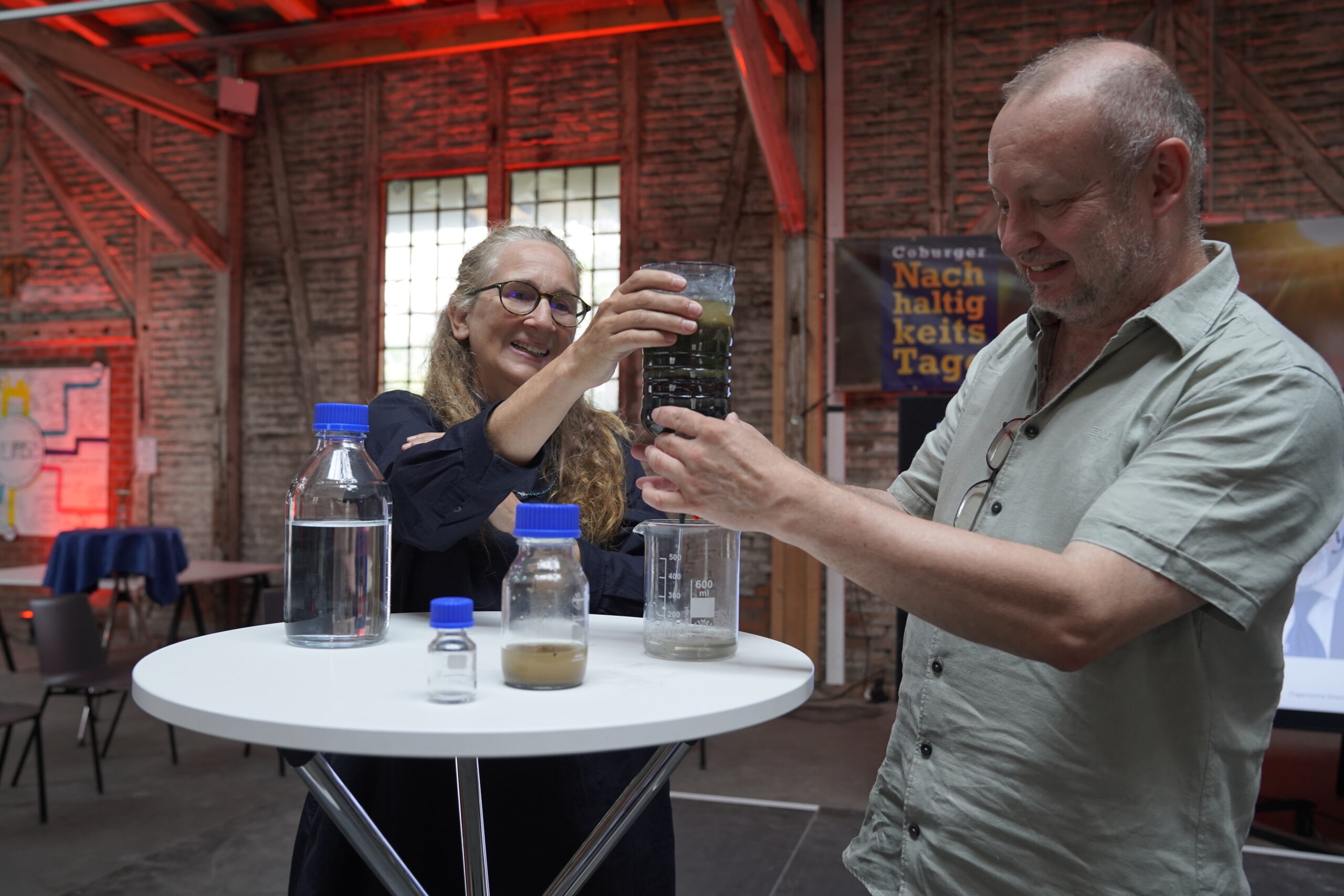 Zwei Menschen stehen an einem runden Tisch mit Flaschen und Gläsern mit Wasserproben. Lächelnd untersuchen sie ein Glas mit dunkler Flüssigkeit in einem Industrieraum mit Backsteinwänden und roter Beleuchtung, der den Geist der Nachhaltigkeitstage 2025 in Coburg einfängt. at Hochschule Coburg