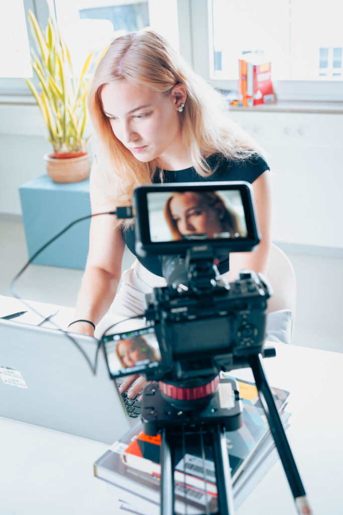Eine Frau mit blondem Haar arbeitet an ihrem Schreibtisch an einem Laptop und filmt sich selbst für ein Projekt der Wirtschaftsinformatik 2.0. Auf dem Kamerabildschirm ist ihr Gesicht zu sehen, eine Topfpflanze und Bücher bilden den studierenden Hintergrund. at Hochschule Coburg