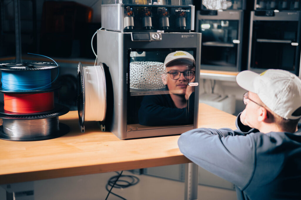Eine Person mit Brille und Mütze schaut nachdenklich auf einen 3D-Drucker, der auf einem Holztisch steht, umgeben von Spulen mit Filamenten - eine Szene, die die Innovation und die analytische Denkweise der Wirtschaftsinformatik widerspiegelt. at Hochschule Coburg