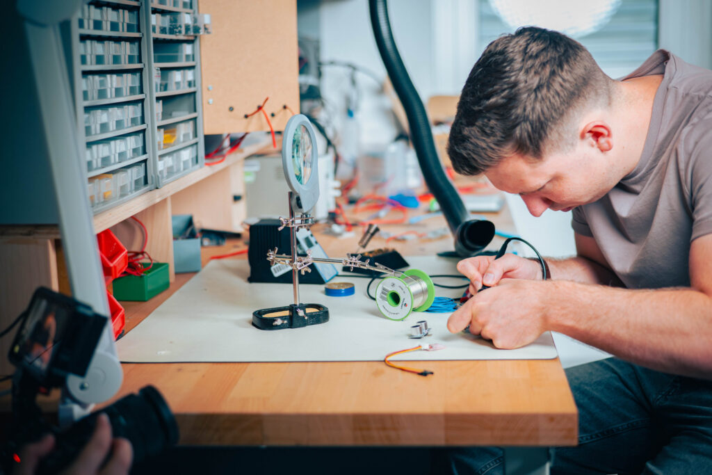 Ein Mann konzentriert sich auf das Löten von elektronischen Bauteilen an einer Werkbank, umgeben von Werkzeugen und Schaltkreisen, und verkörpert damit die praktische Innovation, die oft in Workshops der Wirtschaftsinformatik 2.0 erforscht wird. at Hochschule Coburg