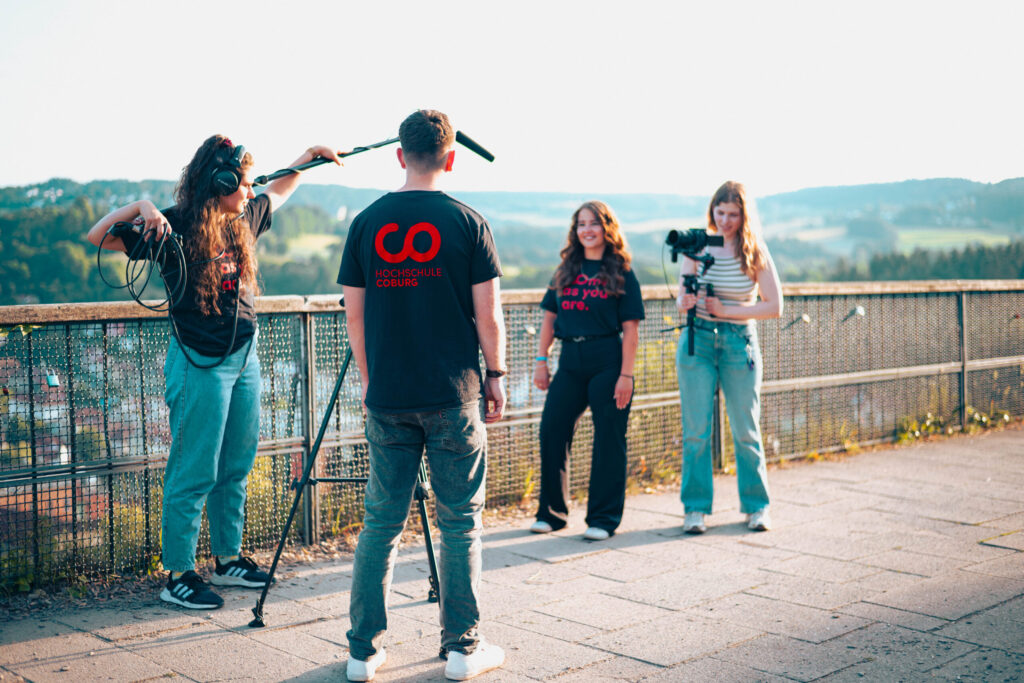 Vier Personen filmen im Freien auf einer gepflasterten Terrasse mit Blick auf die Landschaft. Einer hält ein Boom-Mikrofon in der Hand, ein anderer steht mit dem Rücken zur Kamera, zwei weitere lächeln und fangen den Moment für ein Projekt der Wirtschaftsinformatik 2.0 ein. at Hochschule Coburg