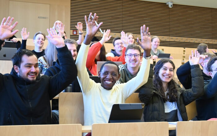 Studierende der Hochschule Coburg sitzen während einer Infoveranstaltung des Orientierungssemesters in einem belebten Hörsaal, lächeln und heben enthusiastisch die Hände. Laptops und Tablets auf den Tischen unterstreichen ihre engagierte Teilnahme. at Hochschule Coburg