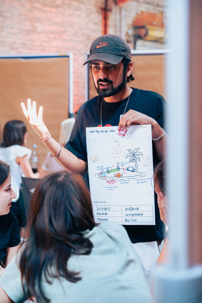 Ein Mann mit schwarzer Mütze und T-Shirt hält ein Plakat mit bunten Zeichnungen und Texten über Betriebswirtschaft hoch und spricht zu einer Gruppe von sitzenden Menschen in einem hellen, legeren Innenraum. at Hochschule Coburg