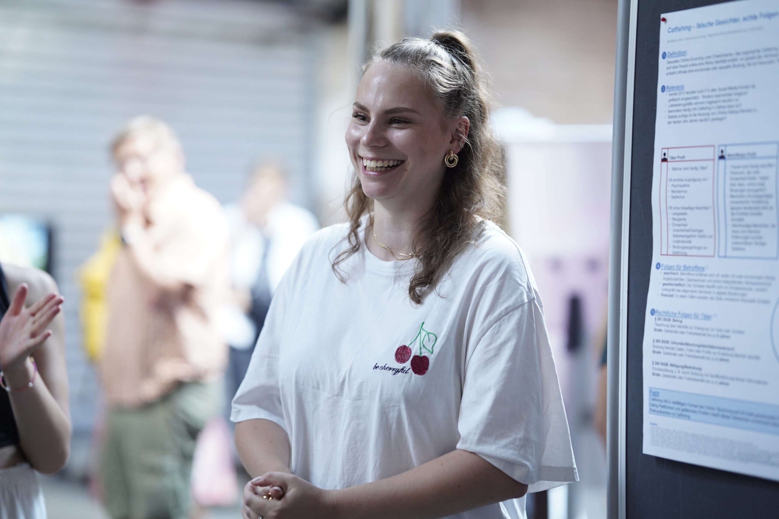 Eine lächelnde junge Frau mit langen Haaren und weißem T-Shirt steht in einem Innenraum neben einem Präsentationsplakat, hinter ihr sind Menschen und verschwommene Hintergründe zu sehen. at Hochschule Coburg