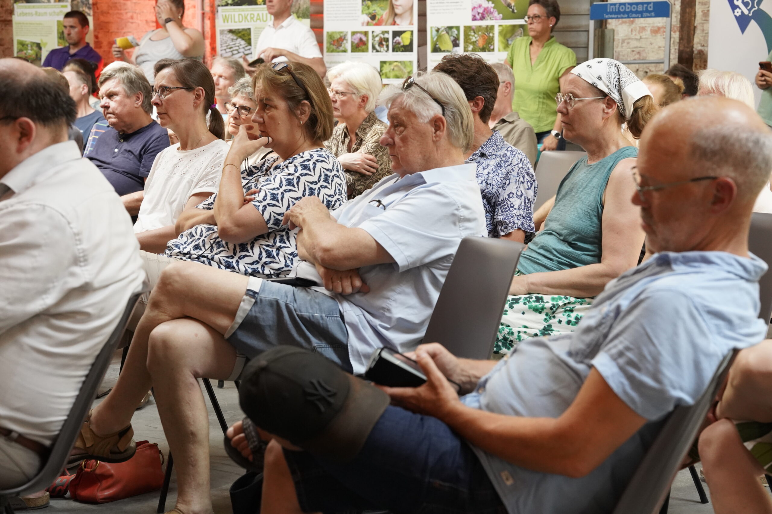 Eine Gruppe Erwachsener sitzt bei einer Indoor-Veranstaltung aufmerksam in Stuhlreihen und hört einem Redner zu. Die meisten Menschen scheinen konzentriert zu sein, einige haben die Arme verschränkt. An der Wand hinter ihnen sind Plakate mit Bildern und Texten zu sehen. at Hochschule Coburg