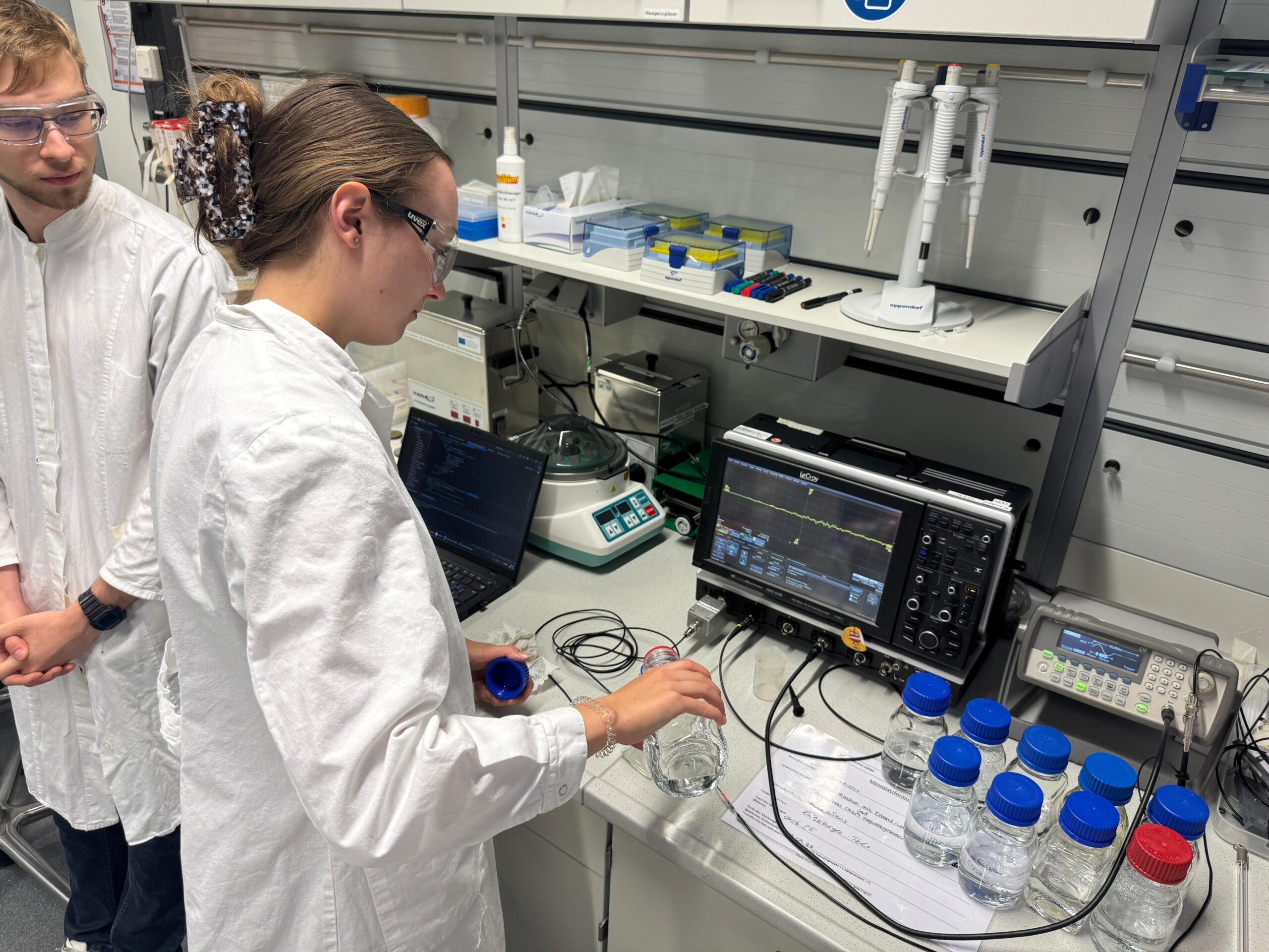 Zwei Personen in Laborkitteln arbeiten an einem Labortisch mit wissenschaftlichen Geräten, Laptops und Flaschen mit blauen Deckeln, die Forschung in der Praxis demonstrieren. Einer stellt ein Gerät ein, das über Kabel mit den Flaschen verbunden ist, während der andere aufmerksam zusieht. at Hochschule Coburg