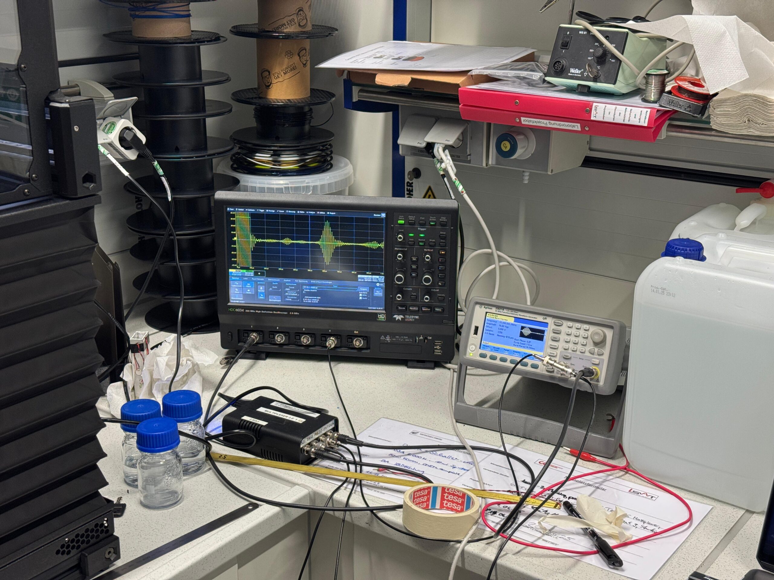 Ein Laborarbeitsplatz mit elektronischen Testgeräten für Sensorik Forschung, darunter ein Oszilloskop, das Wellenformen anzeigt, verschiedene Kabel, Laborflaschen, Dokumente, Klebeband und andere Werkzeuge, die auf einer unordentlichen Arbeitsplatte verstreut sind. at Hochschule Coburg