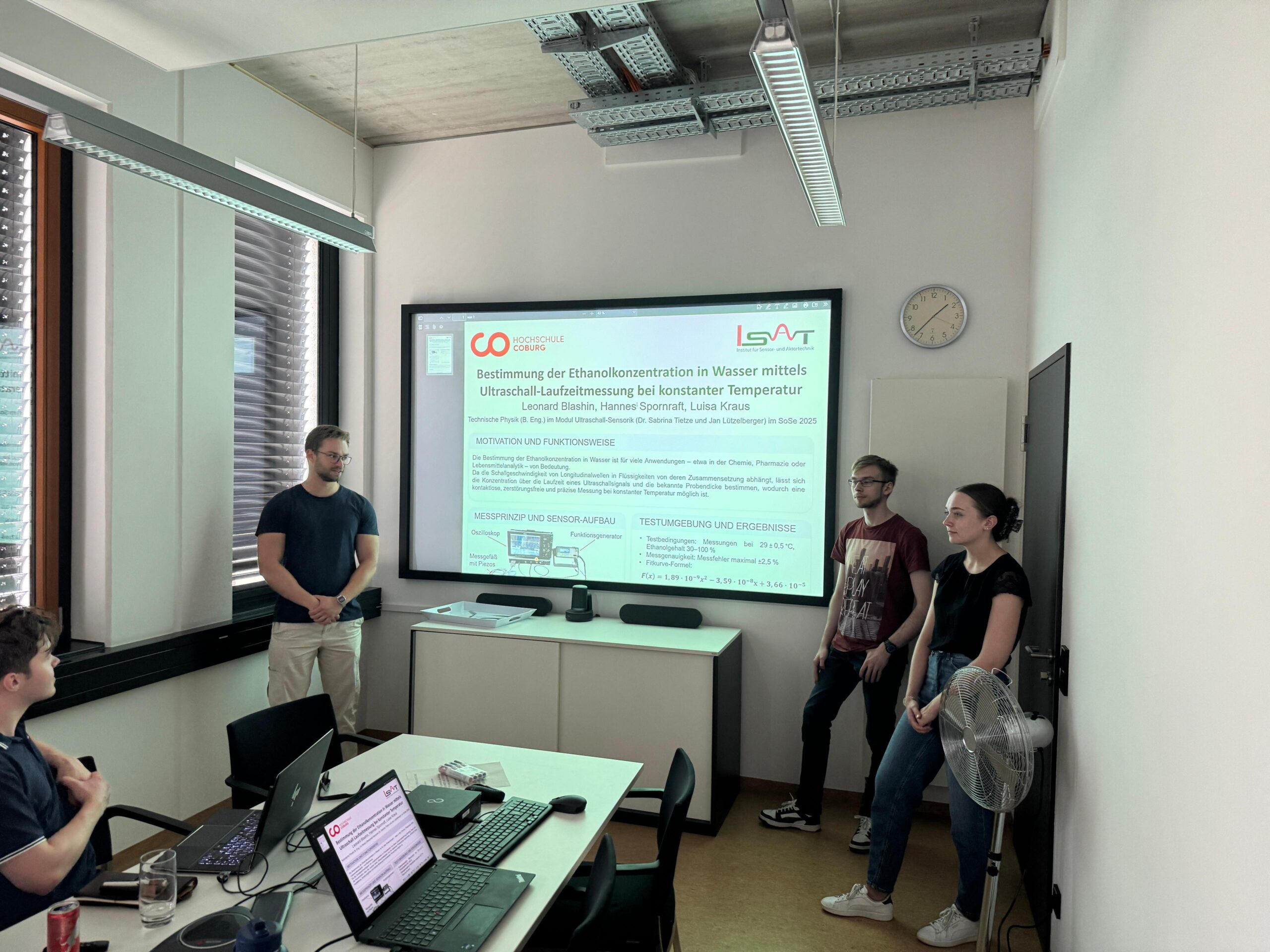 Vier Personen stehen vorne in einem Klassenzimmer und halten einen Vortrag; eine große Folie über Ethanolbestimmung wird projiziert, auf der Forschung und Praxis hervorgehoben werden, während Laptops, Papiere und eine Kaffeetasse auf dem Tisch im Vordergrund verstreut sind. at Hochschule Coburg