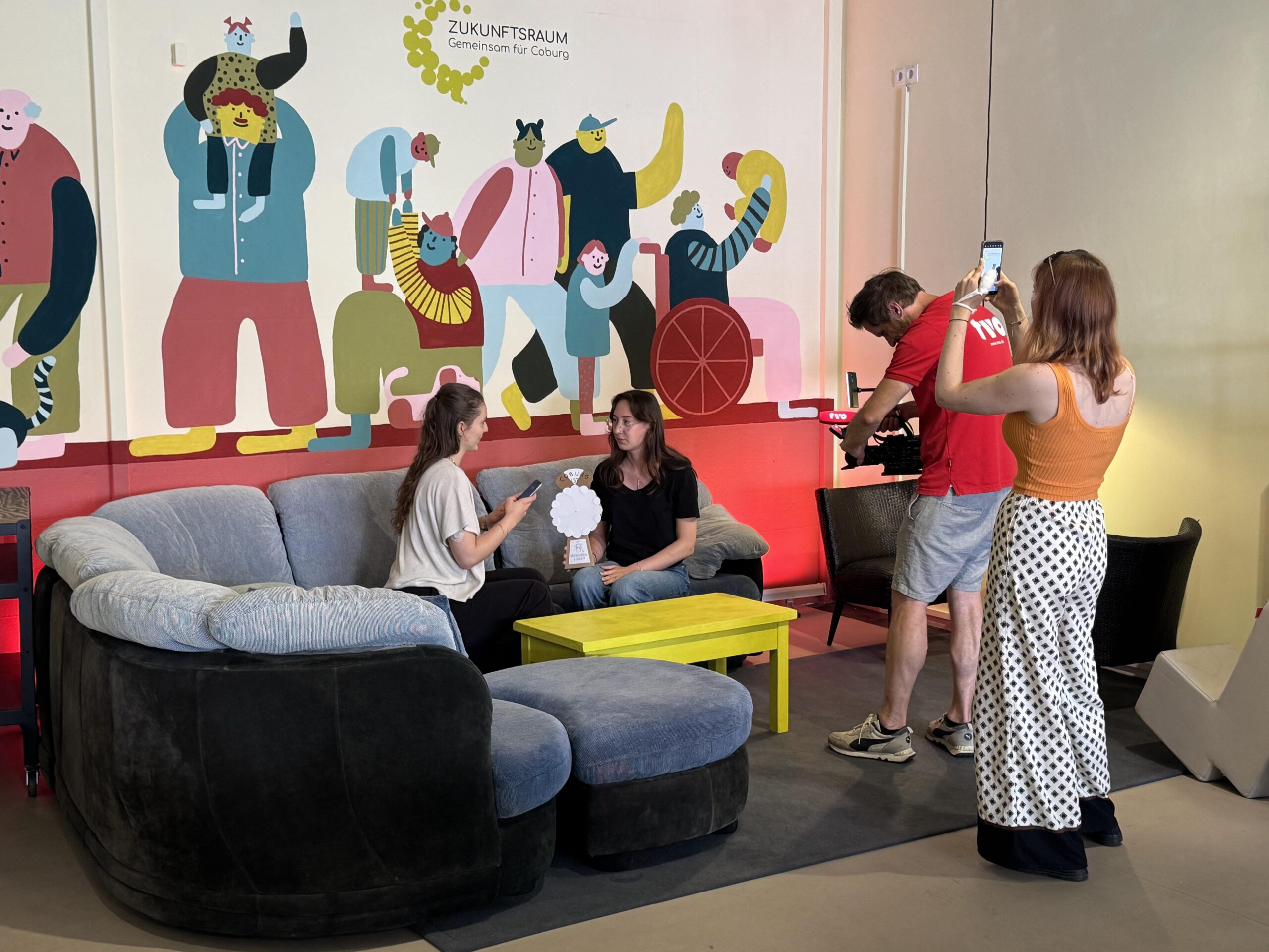 Zwei Frauen sitzen und unterhalten sich auf einer grauen Couch vor einem bunten Wandgemälde, während zwei Personen mit Kameras sie aufnehmen und fotografieren. Vor der Couch steht ein gelber Tisch, und die Umgebung wirkt informell und kreativ. at Hochschule Coburg