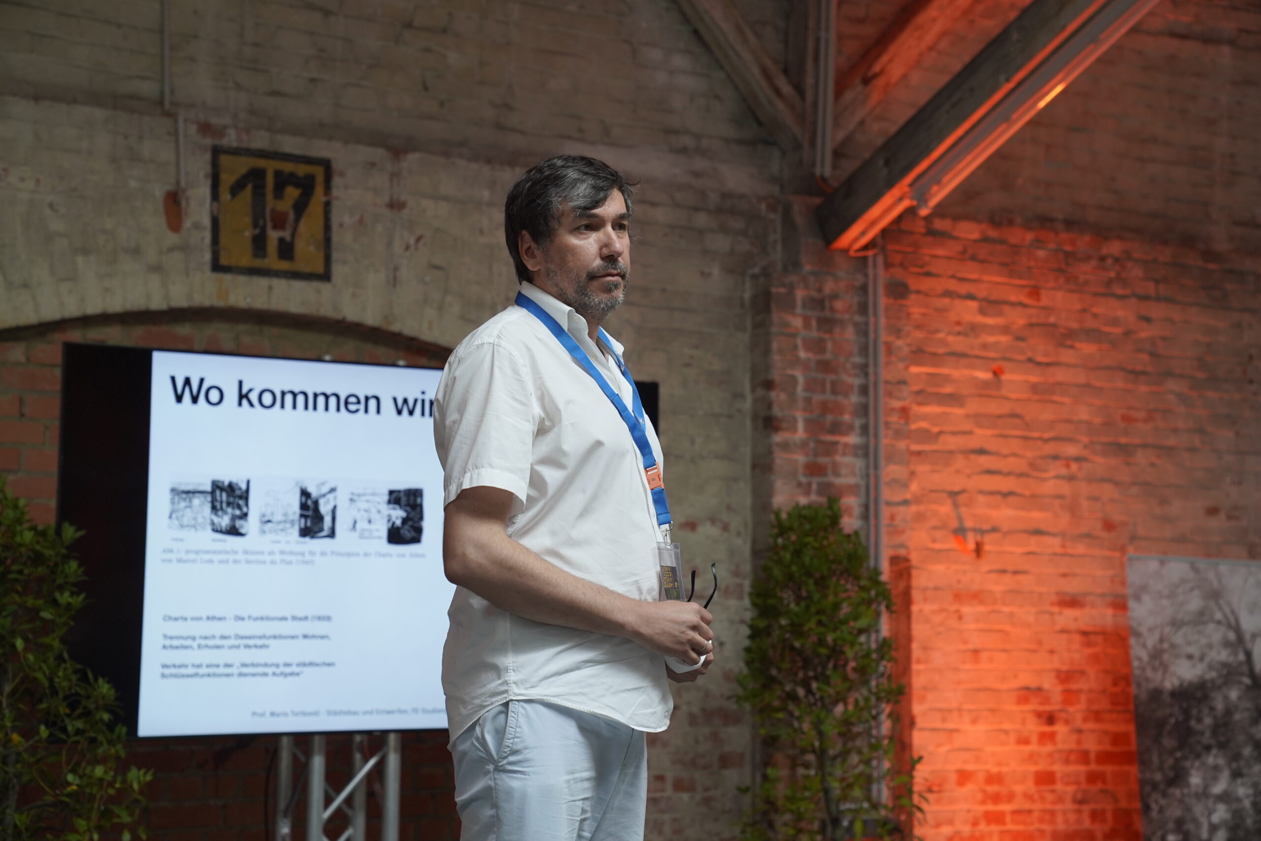 Ein Mann in einem weißen Hemd steht vor einem Präsentationsbildschirm mit deutschem Text in einem Industrieraum mit freiliegenden Backsteinwänden und orangefarbenem Licht. Auf dem Bildschirm erscheinen "Wo kommen wi" und historische Fotos. at Hochschule Coburg