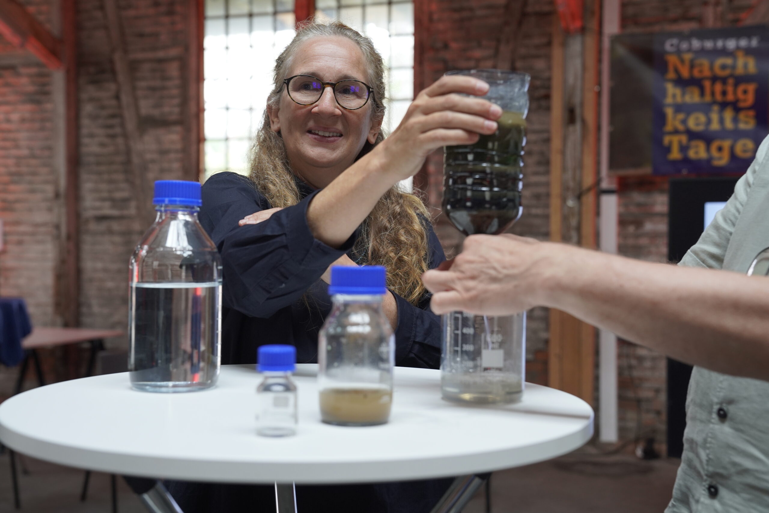 Eine Frau mit langen lockigen Haaren und Brille lächelt, während sie eine Flasche mit dunkler Flüssigkeit an einem runden Tisch hält, an dem verschiedene Flaschen mit Flüssigkeiten ausgestellt sind. Die Hand einer anderen Person greift hinein, und im Hintergrund ist ein "Nachhaltigkeitstage"-Schild zu sehen. at Hochschule Coburg