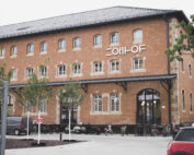 Ein großes Backsteingebäude mit vielen Fenstern und einem Schild mit der Aufschrift "COIIH-OF" an der Fassade. Am Eingang sind Fahrräder abgestellt, und auf dem Parkplatz stehen ein paar Autos und Motorräder. Die Vorderseite ist von Bäumen gesäumt. at Hochschule Coburg