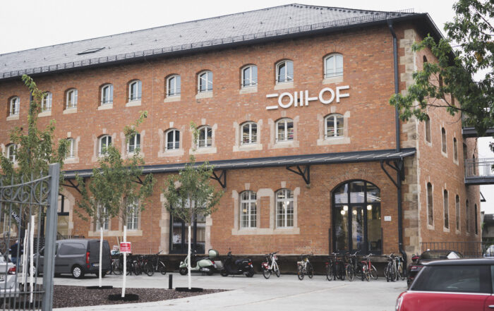 Ein großes Backsteingebäude mit vielen Fenstern und einem Schild mit der Aufschrift "COIIH-OF" an der Fassade. Am Eingang sind Fahrräder abgestellt, und auf dem Parkplatz stehen ein paar Autos und Motorräder. Die Vorderseite ist von Bäumen gesäumt. at Hochschule Coburg