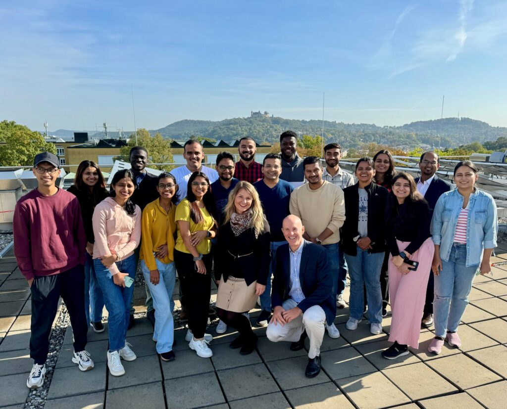 Eine Gruppe von 18 Personen steht lächelnd auf einem Dach mit Bergen und einer Burg im Hintergrund und genießt einen sonnigen Tag unter einem klaren blauen Himmel. at Hochschule Coburg