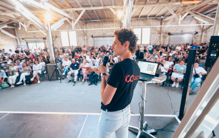 Eine Person spricht auf der Bühne vor einem großen Publikum in ein Mikrofon und bietet ein echtes Live-Erlebnis in einem großen Saal mit freiliegenden Balken. Der Laptop des Sprechers liegt aufgeklappt auf einem hohen Tisch neben ihm. at Hochschule Coburg