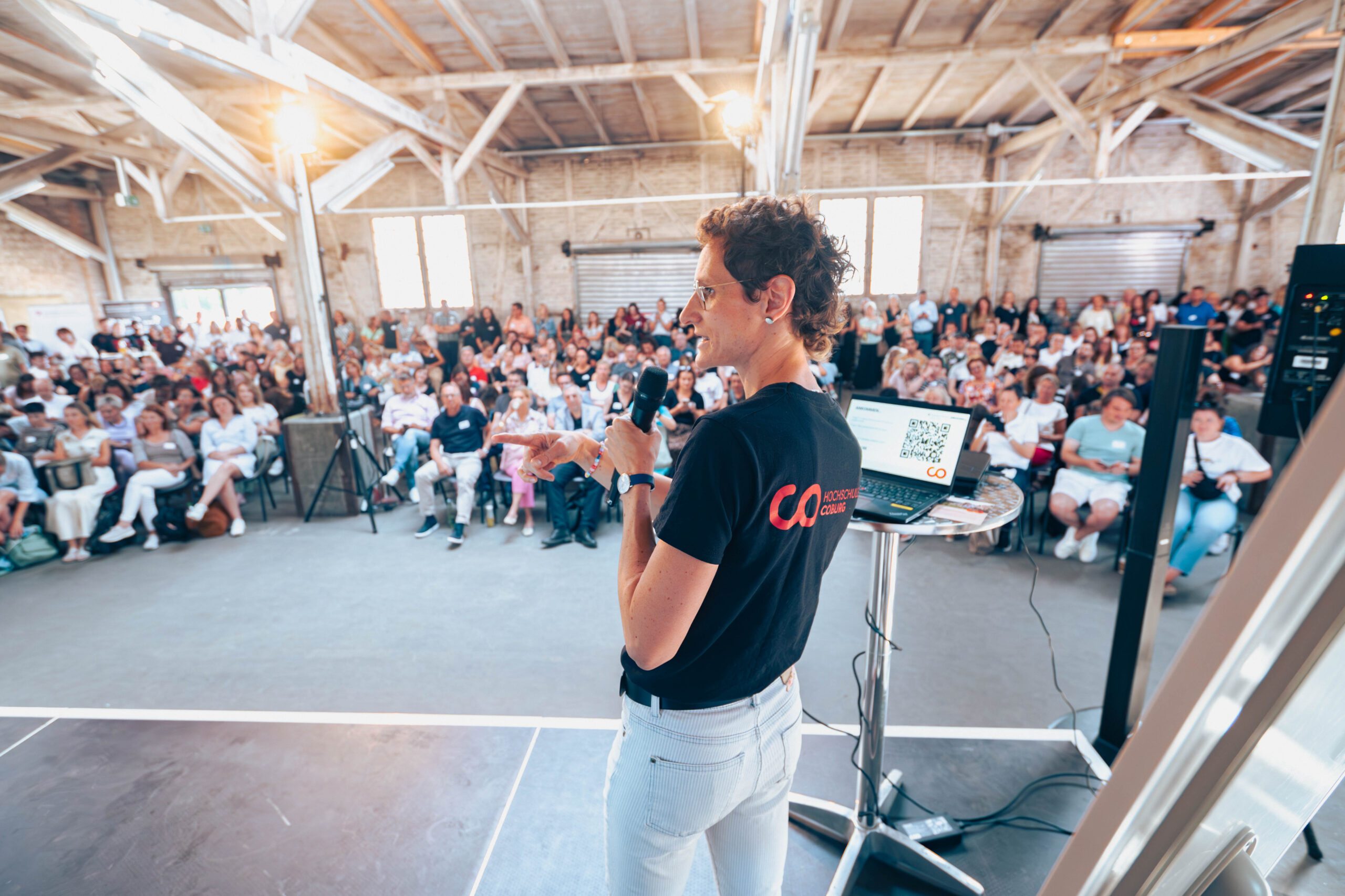 Eine Person spricht auf der Bühne vor einem großen Publikum in ein Mikrofon und bietet ein echtes Live-Erlebnis in einem großen Saal mit freiliegenden Balken. Der Laptop des Sprechers liegt aufgeklappt auf einem hohen Tisch neben ihm. at Hochschule Coburg