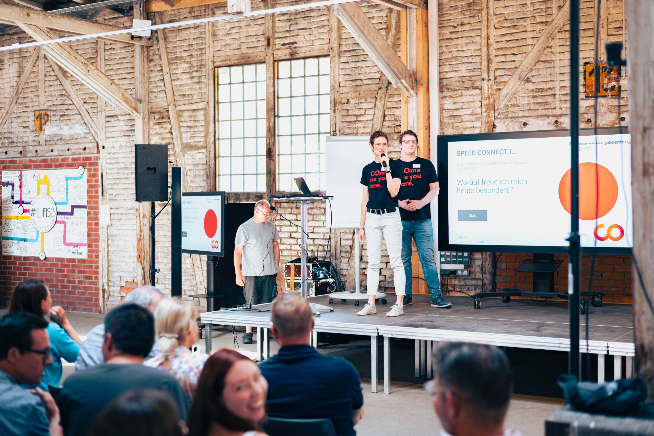 Zwei Männer stehen auf einer Bühne und sprechen in ein Mikrofon bei einer Indoor-Veranstaltung und bieten ein Live-Erlebnis mit großen Bildschirmen hinter ihnen, die Texte und Grafiken anzeigen. Das Publikum, darunter viele Studierende, sitzt der Bühne gegenüber in einem industriell anmutenden Raum. at Hochschule Coburg