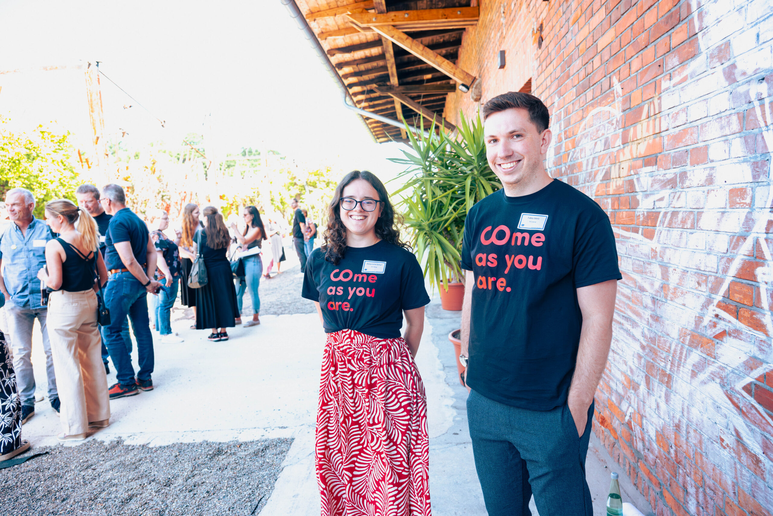 Zwei Studierende in schwarzen "Come as you are."-T-Shirts stehen lächelnd vor einem Backsteingebäude; im Hintergrund ist eine Gruppe von Menschen versammelt, die an einem sonnigen Tag ein lebendiges Live-Erlebnis genießen. at Hochschule Coburg