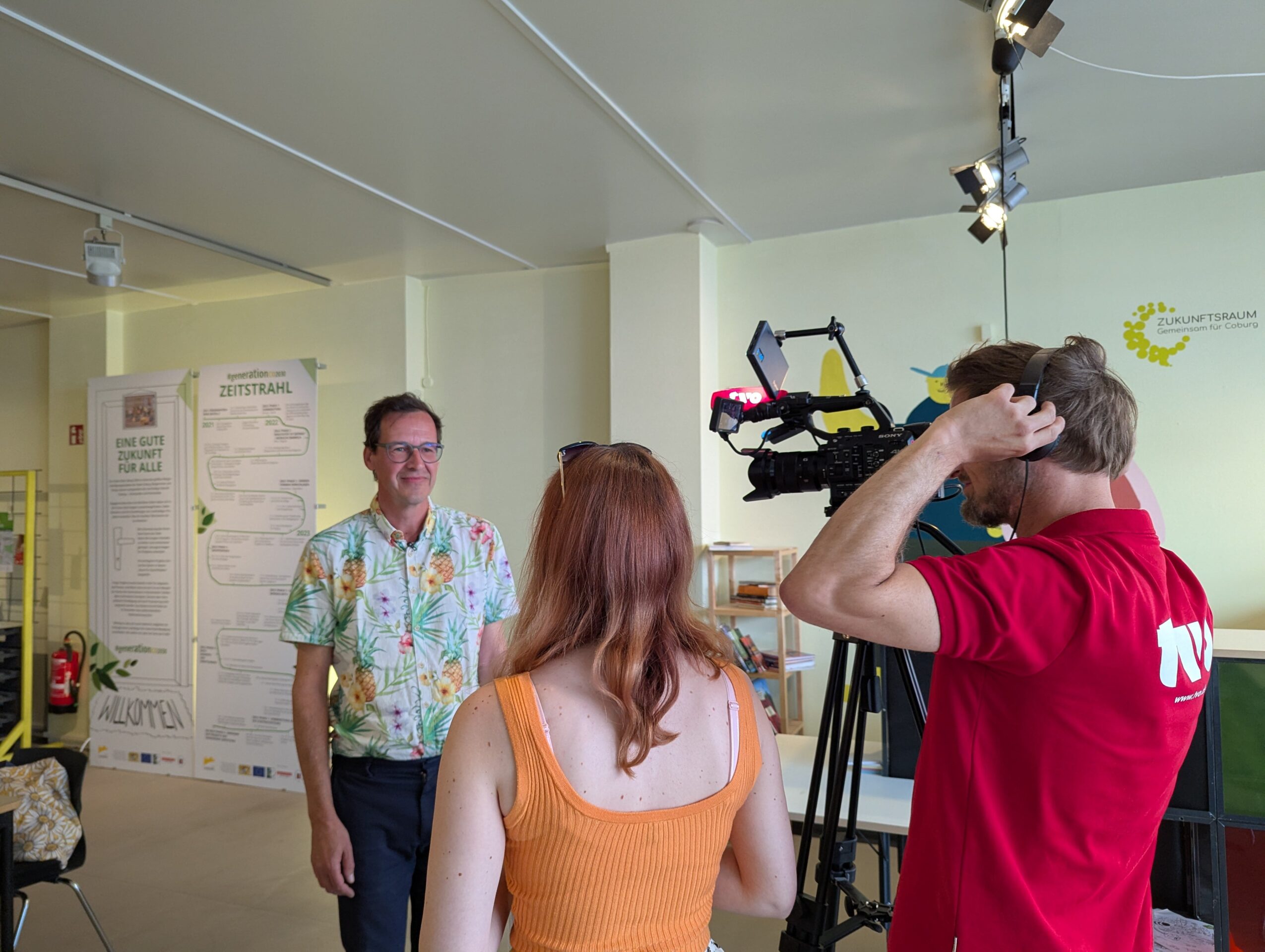 Ein Mann in einem geblümten Hemd wird von einer Frau interviewt, während ein Kameramann die beiden in einem Innenraum filmt. Im Hintergrund sind Plakate und Informationsdisplays zu sehen. at Hochschule Coburg