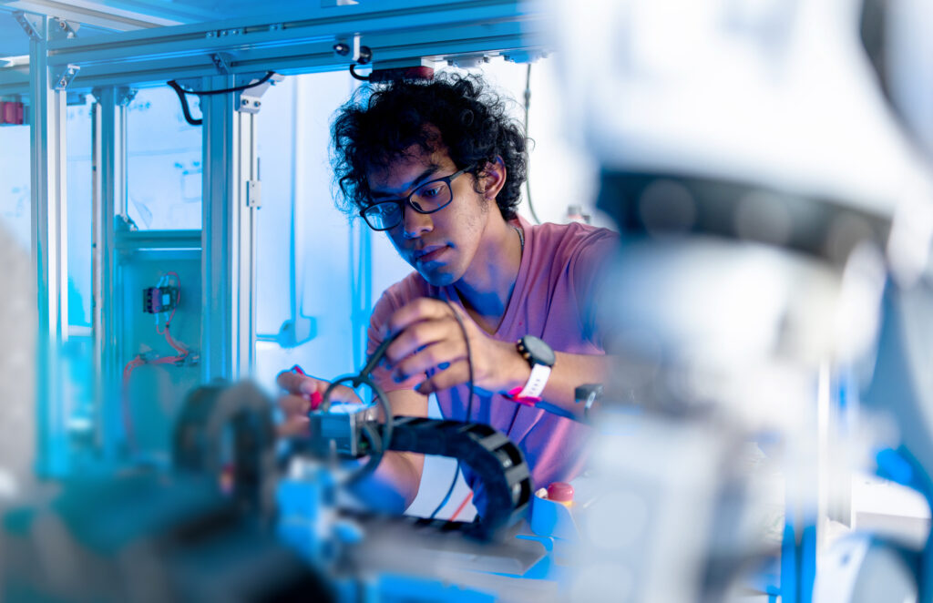 Eine Person mit Brille und rosa Hemd arbeitet in einem Hightech-Labor mit blauer Beleuchtung an einem Robotergerät und stellt mit Werkzeugen Bauteile ein - eine inspirierende Szene an der Schnittstelle von Technik und Informatik. at Hochschule Coburg