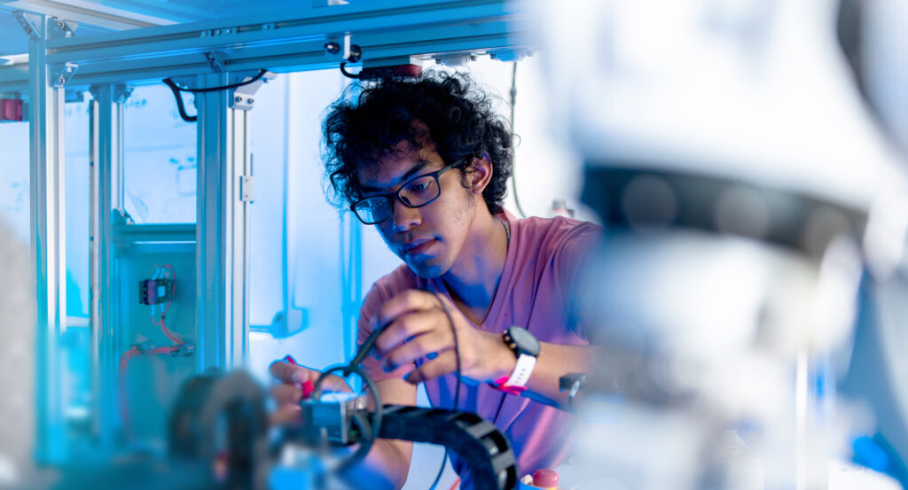 A person wearing glasses and a pink shirt works intently with wires and tools on a robotic device in a brightly lit, high-tech engineering laboratory environment. at Hochschule Coburg