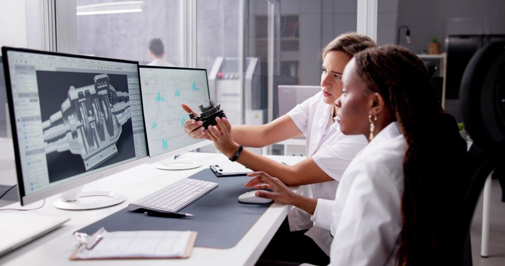 Two women sit at a desk in an engineering office, analyzing a part while looking at 3D technical schematics on a computer screen. One holds a mechanical component, and both are focused intently on the monitor. at Hochschule Coburg