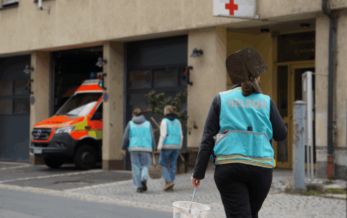 Eine Person in einer blauen "HELPON"-Weste, die zu einem Team der Katastrophenhilfe gehört, trägt einen weißen Eimer und eine Schaufel in der Nähe einer Straße, während zwei andere Personen in ähnlichen Westen auf einen Rettungswagen vor einem Gebäude mit einem roten Kreuz zugehen. at Hochschule Coburg