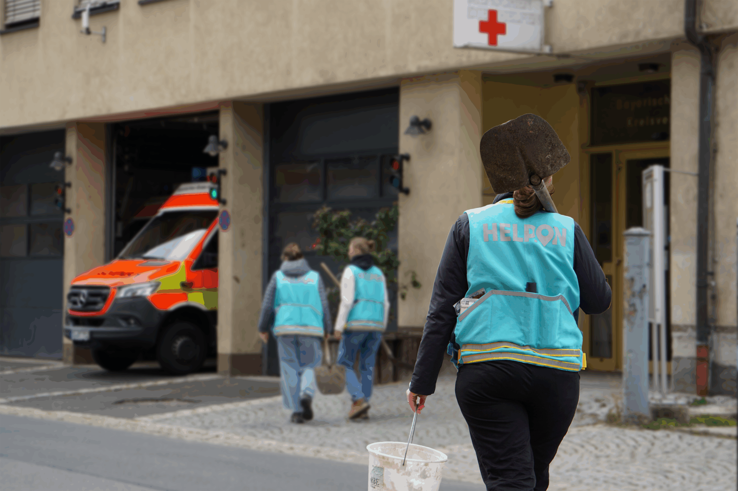 Eine Person in einer blauen "HELPON"-Weste, die zu einem Team der Katastrophenhilfe gehört, trägt einen weißen Eimer und eine Schaufel in der Nähe einer Straße, während zwei andere Personen in ähnlichen Westen auf einen Rettungswagen vor einem Gebäude mit einem roten Kreuz zugehen. at Hochschule Coburg