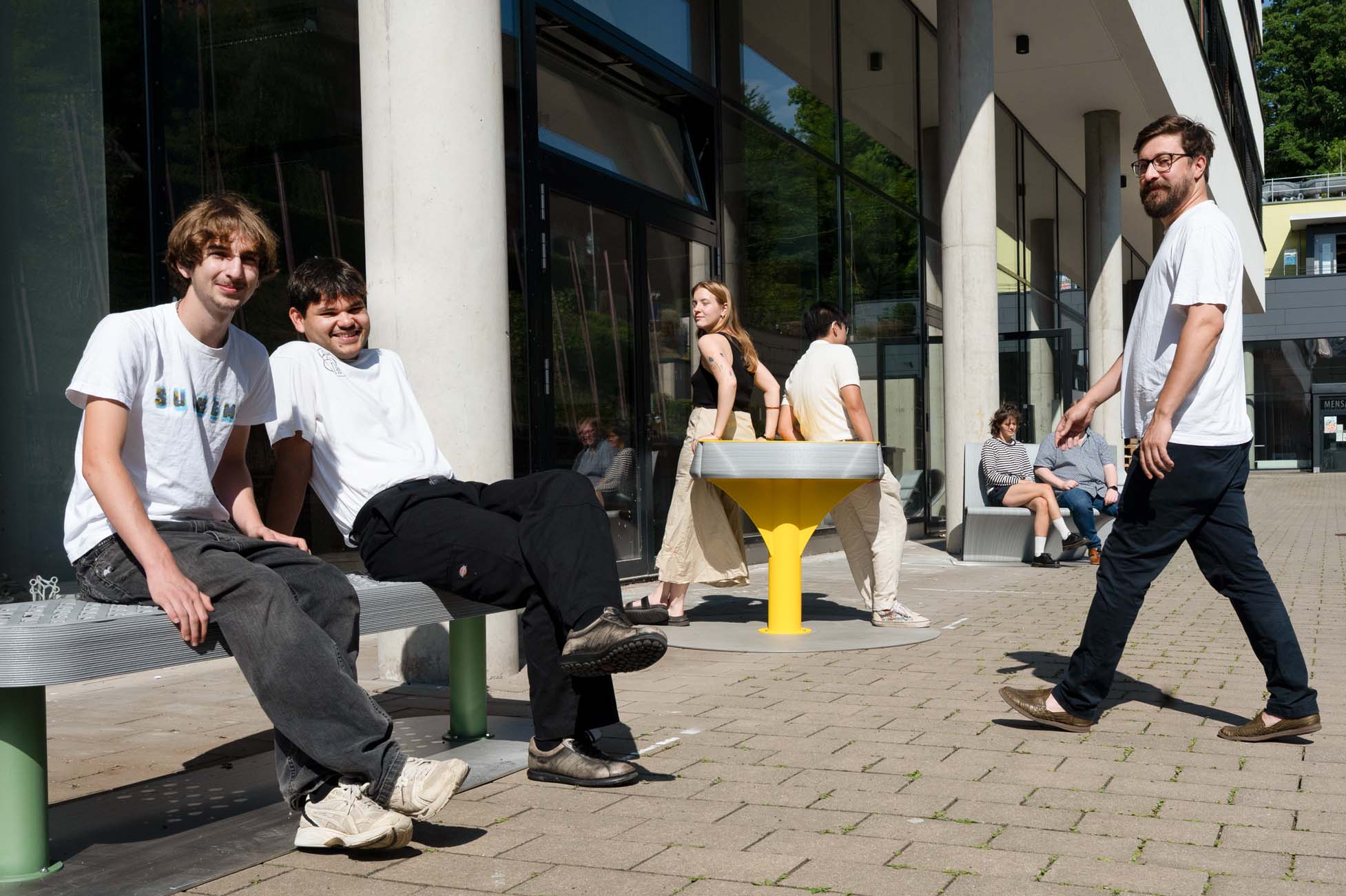 Mehrere Personen entspannen sich vor einem modernen Gebäude; zwei junge Männer sitzen auf einer Bank im Vordergrund, während sich andere um Bänke und einen gelben Tisch versammeln. Die Atmosphäre ist sonnig und entspannt - ein automatisch gespeicherter Schnappschuss, wie ein automatisch gespeicherter Entwurf. at Hochschule Coburg