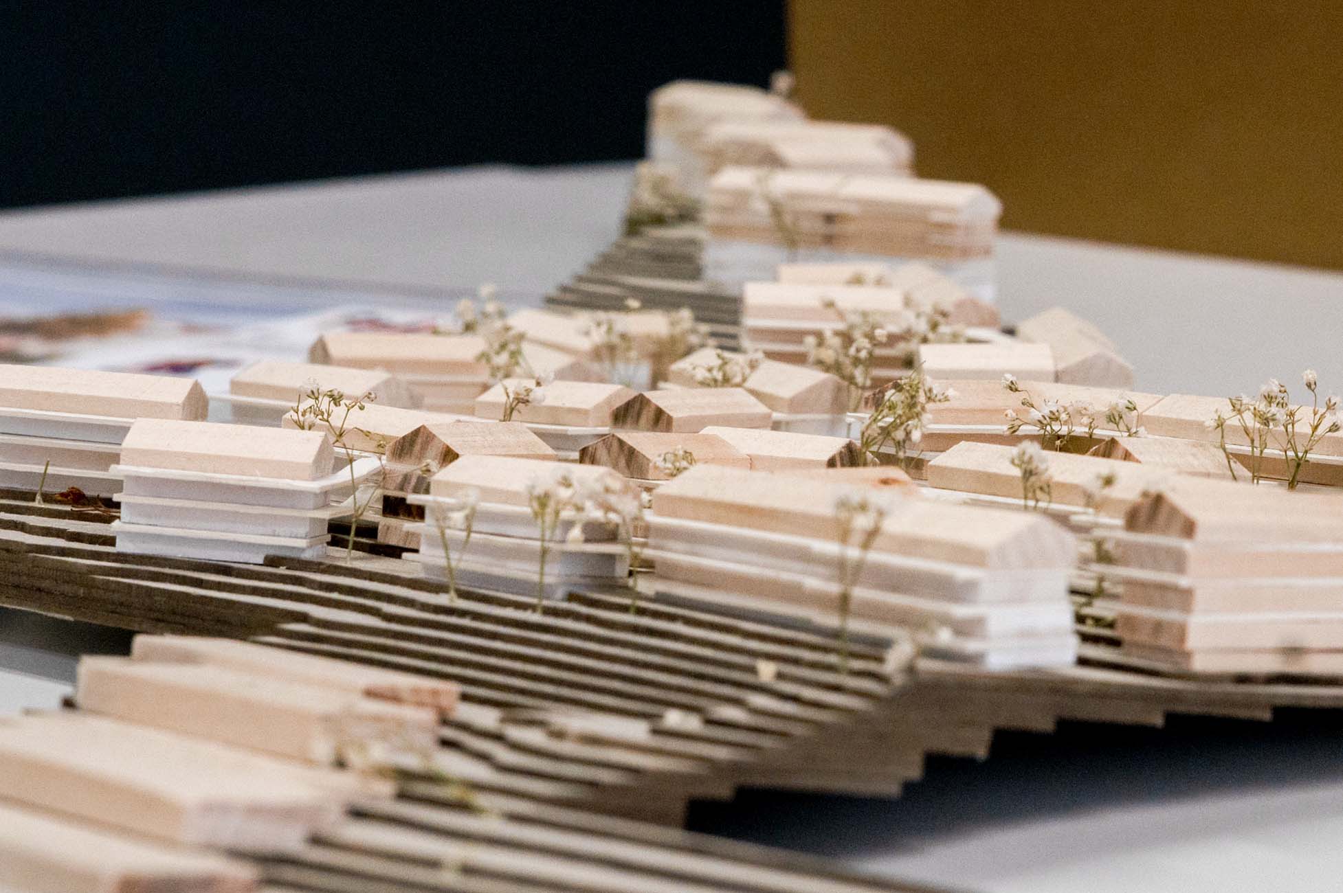 Eine Nahaufnahme eines hölzernen Architekturmodells mit der Aufschrift "Automatisch gespeicherter Entwurf" zeigt kleine, häuserähnliche Blockstrukturen, zwischen denen dünne, getrocknete Pflanzenstängel auf einem geschichteten Holzsockel liegen. at Hochschule Coburg