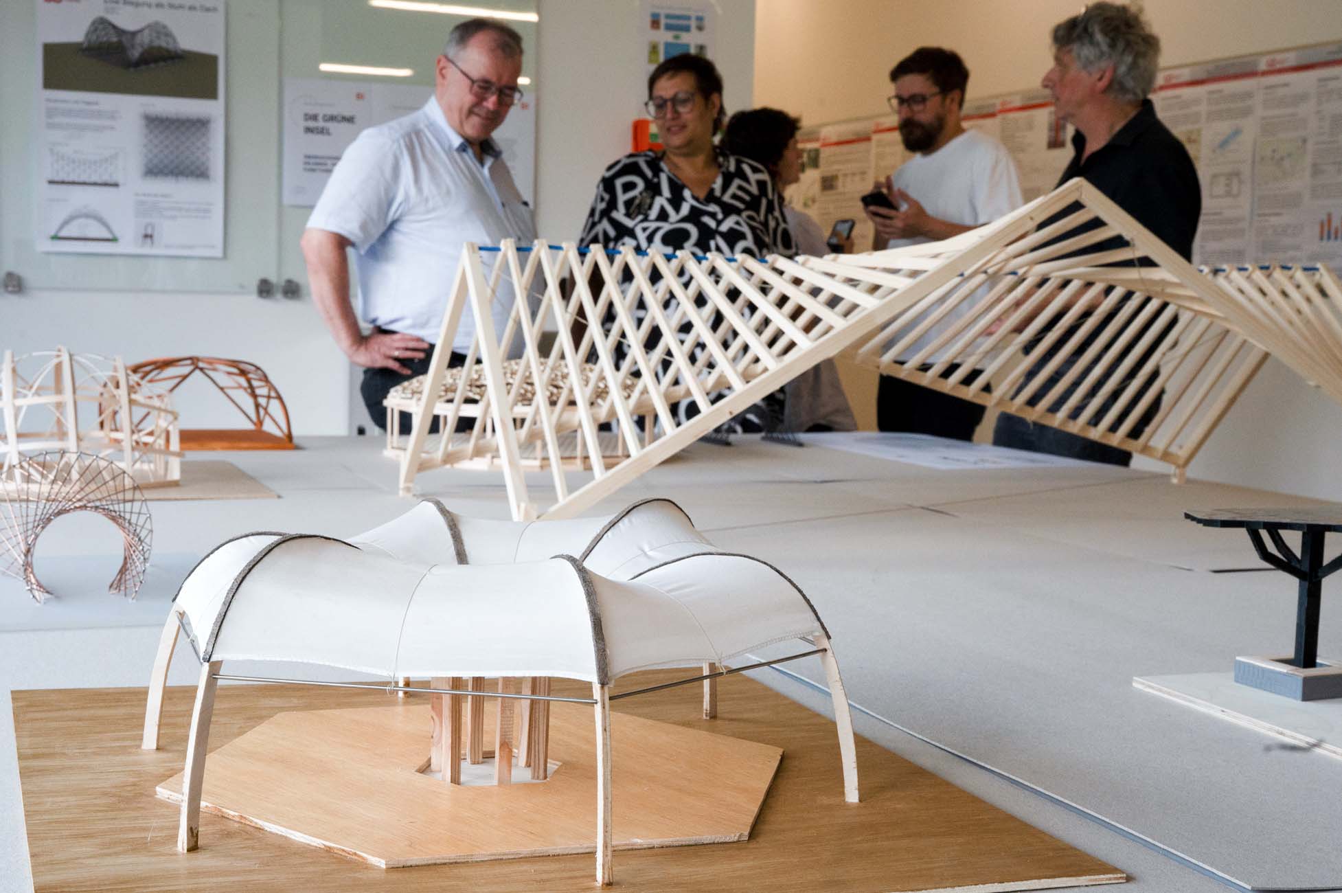 Vier Personen stehen in einem Raum und diskutieren über Architekturmodelle, darunter Holz- und Stoffkonstruktionen. An den Wänden im Hintergrund sind Plakate mit Diagrammen und Text zu sehen, eines mit der Aufschrift "automatisch gespeicherter Entwurf". at Hochschule Coburg