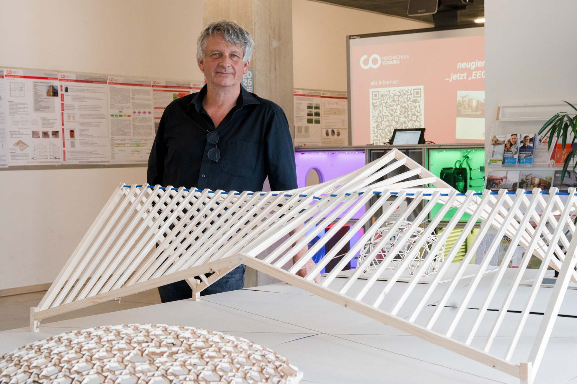 Ein Mann steht hinter einer geometrischen Holzstruktur, die auf einem Tisch in einem modernen Ausstellungsraum ausgestellt ist, während im Hintergrund Informationsplakate und ein digitaler Bildschirm zu sehen sind - ein automatisch gespeicherter Entwurf, der im Mittelpunkt steht. at Hochschule Coburg