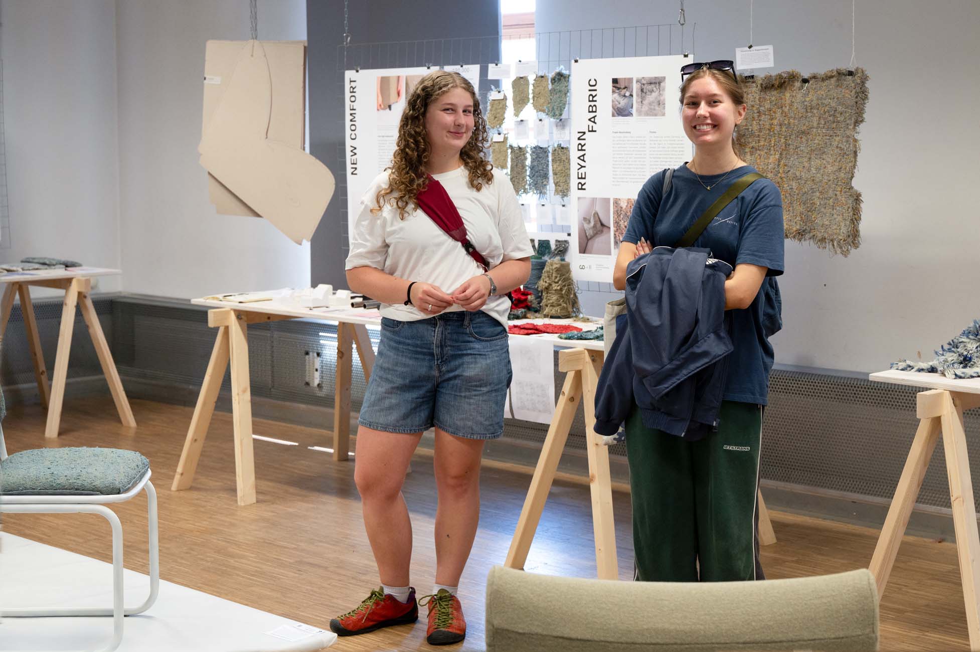 Zwei junge Frauen stehen lächelnd in einem Galerieraum mit Textilkunstauslagen hinter ihnen. Die eine trägt ein weißes T-Shirt und kurze Hosen, die andere ein dunkles T-Shirt und grüne Hosen. Stoffmuster und Informationsplakate sind zu sehen. at Hochschule Coburg