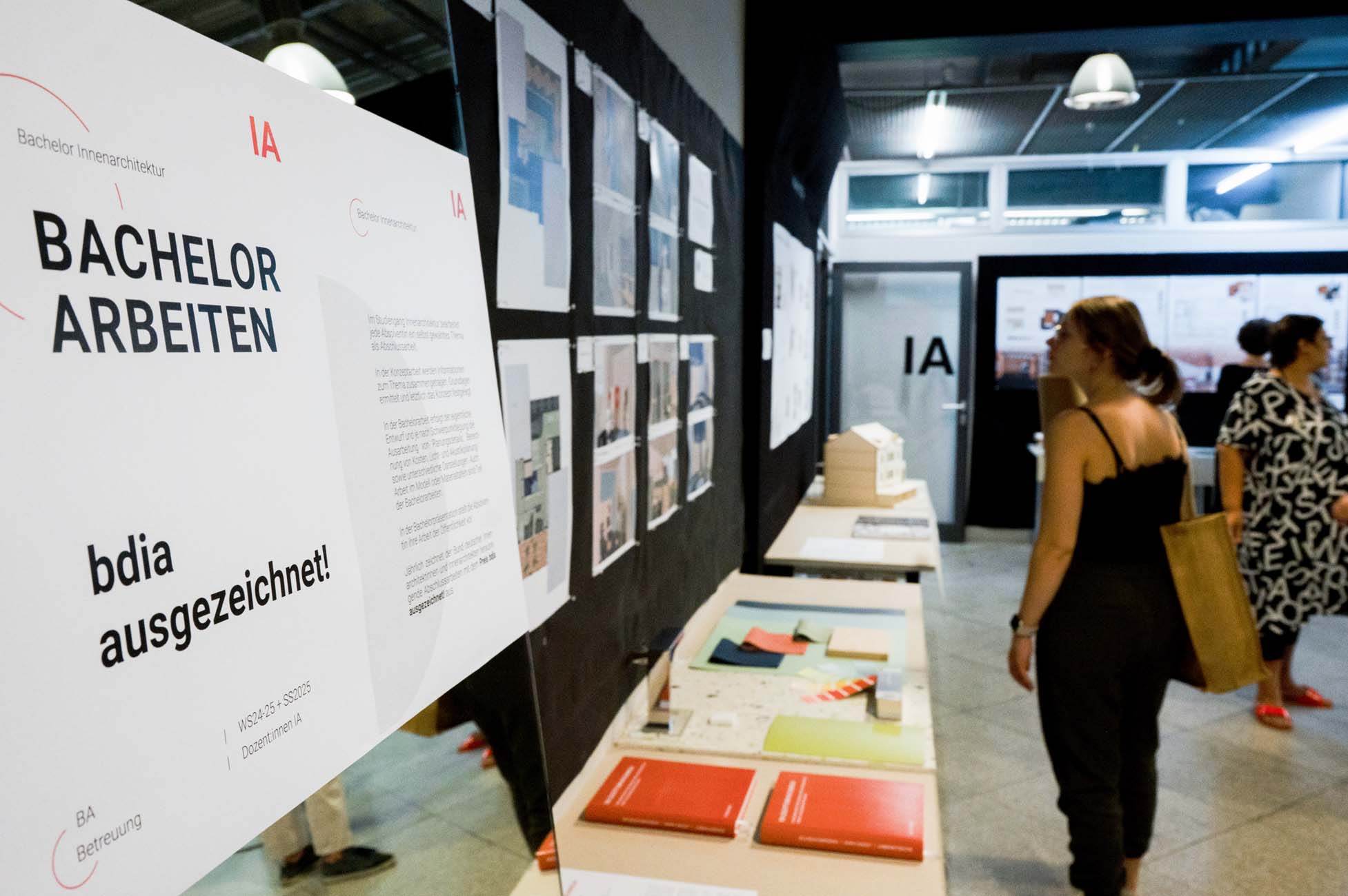 Ausstellungsraum, in dem Bachelor-Arbeiten mit Postern, Broschüren und Architekturmodellen ausgestellt werden; zwei Frauen betrachten die ausgestellten Exponate. Auf einem Schild auf Deutsch steht "Bachelor Arbeiten" und "bdia ausgezeichnet!". at Hochschule Coburg