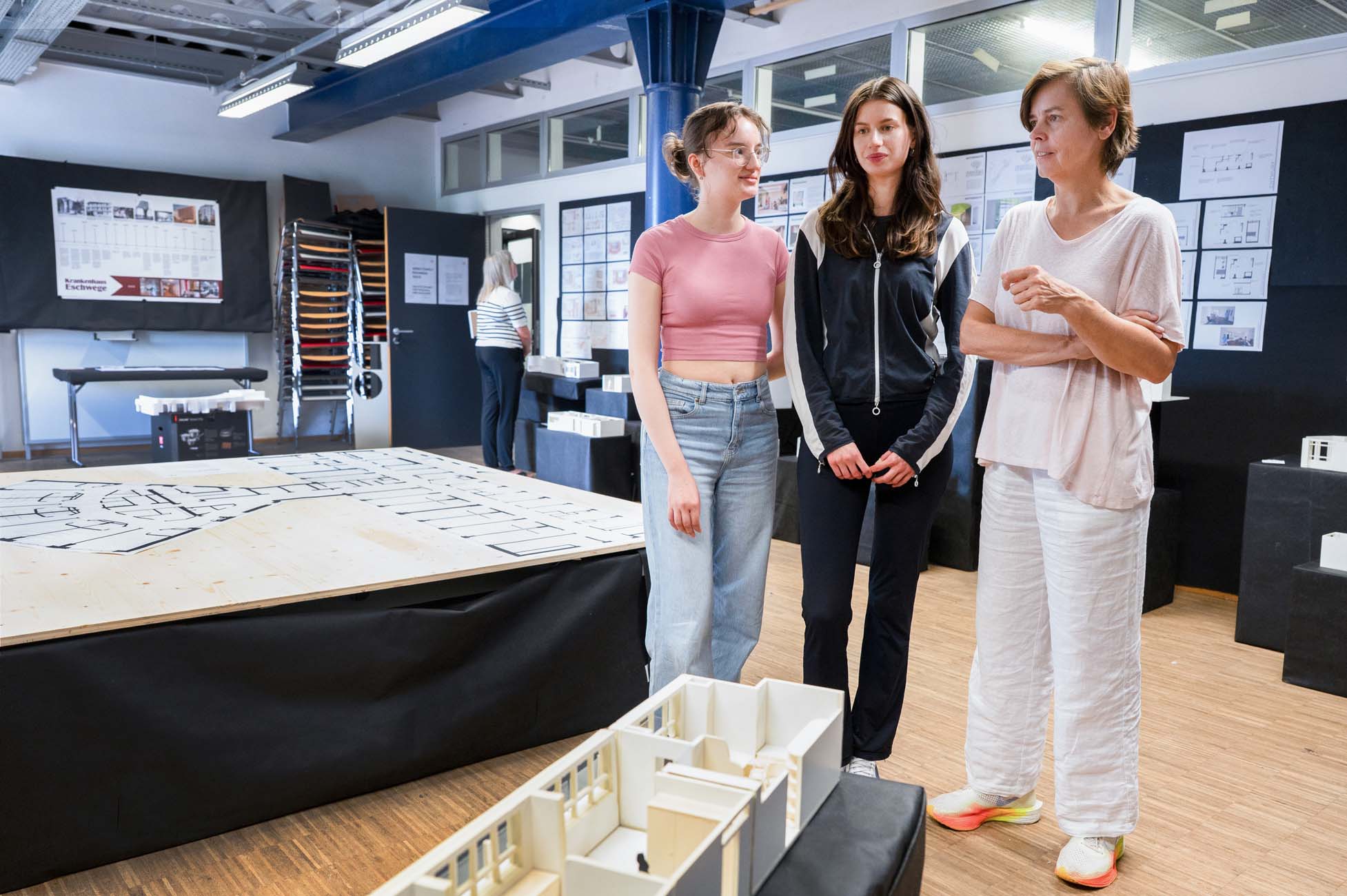Drei Frauen stehen und unterhalten sich in einem Klassenzimmer oder Studio, in dem Architekturmodelle und Grundrisse ausgestellt sind. Eine trägt ein rosafarbenes Crop-Top, eine andere eine dunkle Jacke und die dritte helle Kleidung. Architekturzeichnungen bedecken die Wände. at Hochschule Coburg