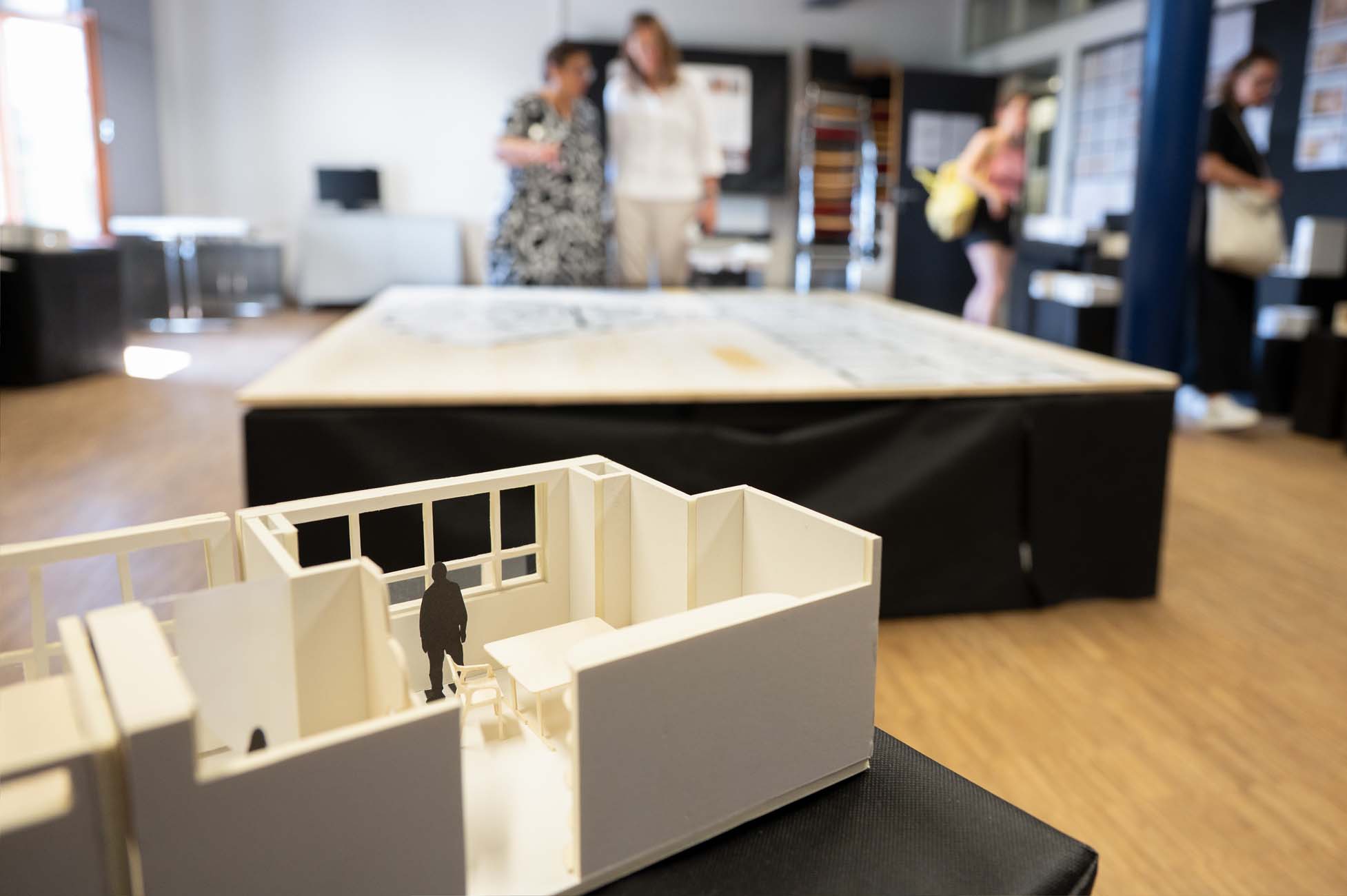 Nahaufnahme eines kleinen architektonischen Modells eines Raums mit einer winzigen Figur darin, die auf einem Tisch steht. Im Hintergrund betrachten vier unscharfe Personen einen großen Grundriss in einem gut beleuchteten Ausstellungsraum. at Hochschule Coburg