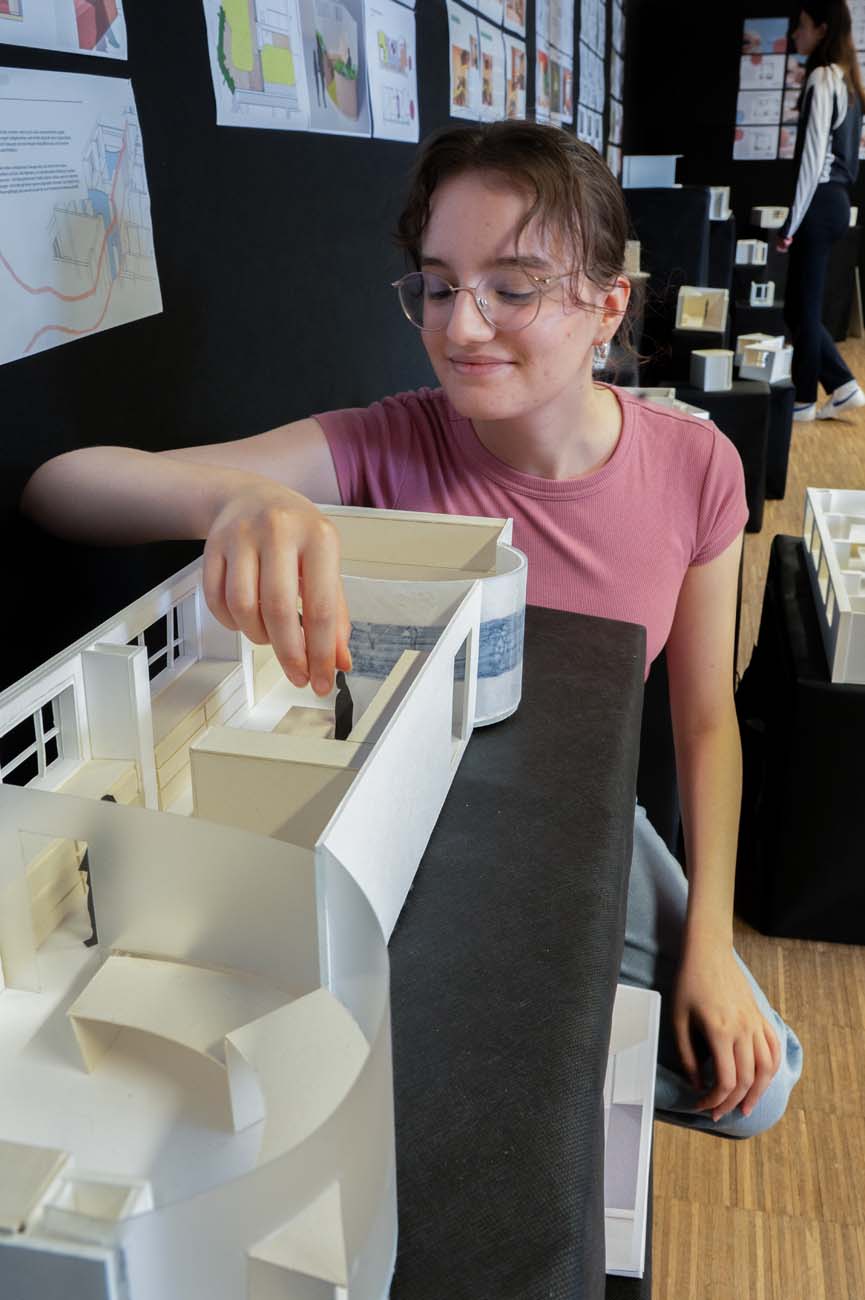 Eine junge Person mit Brille und rosafarbenem Hemd lächelt, während sie eine kleine Figur in ein weißes Architekturmodell stellt, während im Hintergrund Konstruktionspläne und weitere Modelle zu sehen sind. at Hochschule Coburg