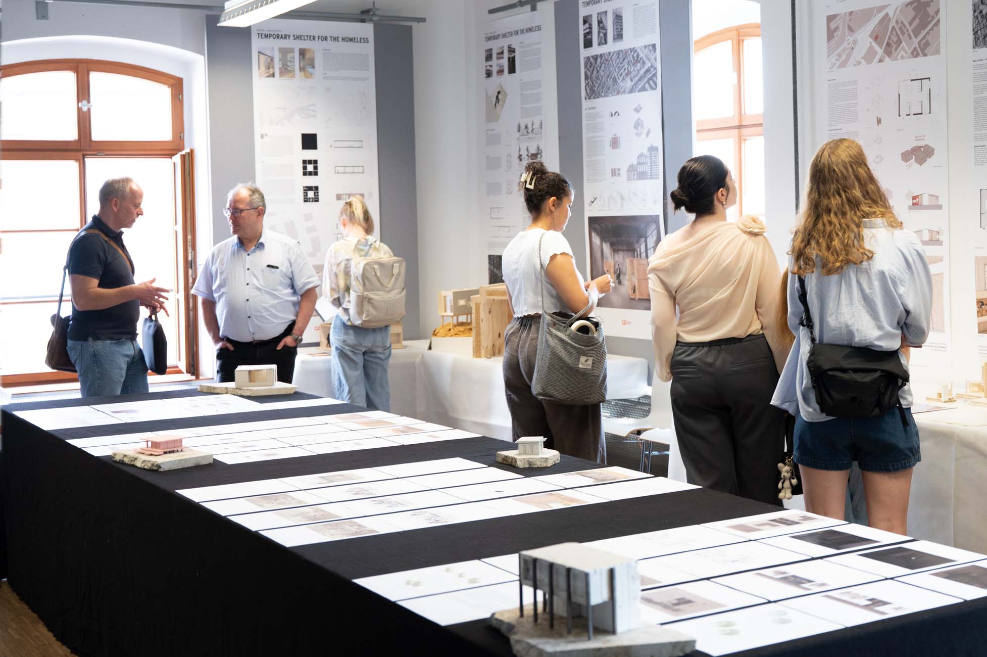 In einem gut beleuchteten Ausstellungsraum werden auf Tischen und an Wänden ausgestellte Architekturmodelle und Entwurfstafeln betrachtet. Einige Besucher kommen ins Gespräch, während andere die Exponate betrachten. at Hochschule Coburg