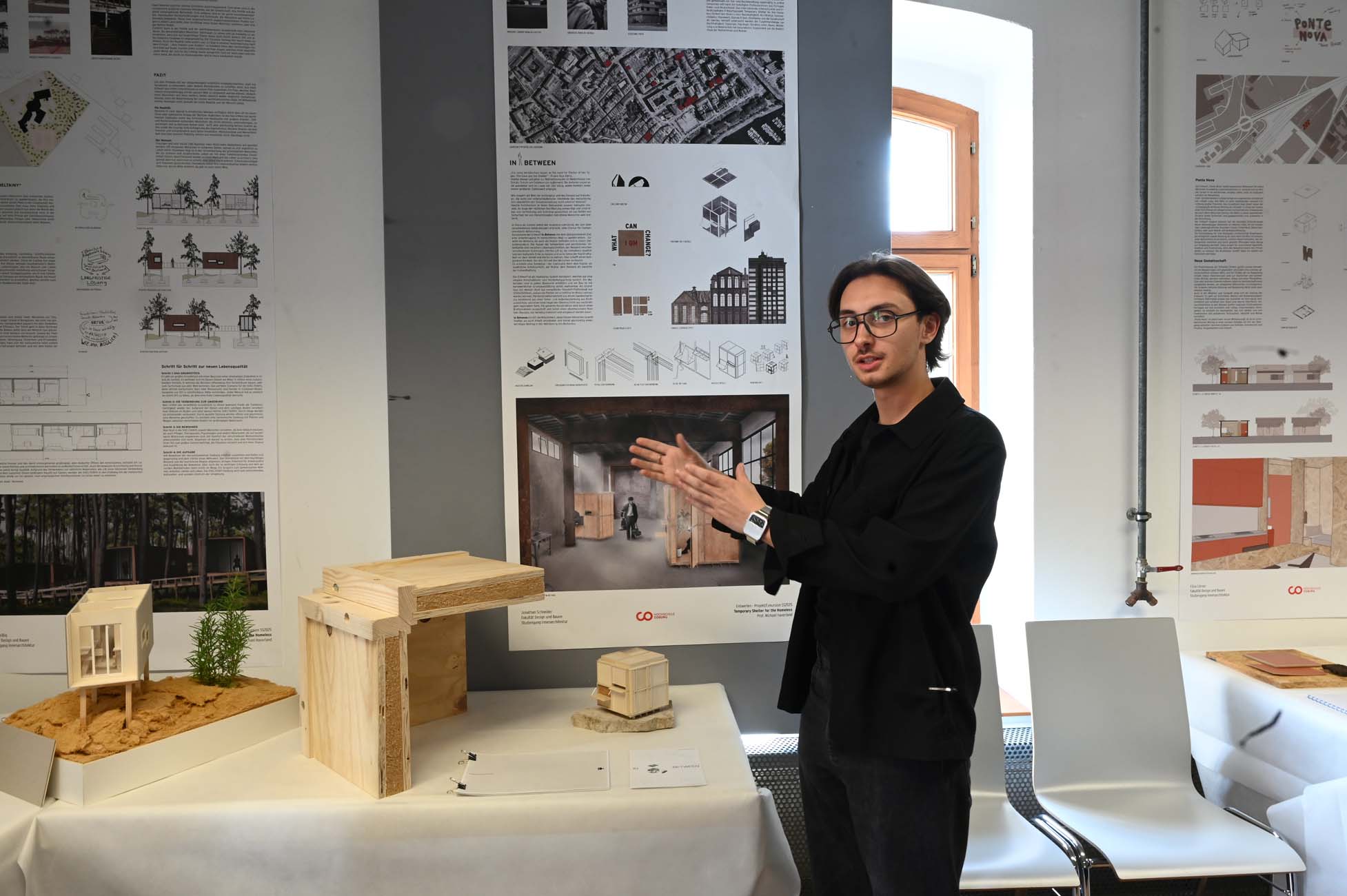 Ein Mann mit Brille gestikuliert in Richtung von Architekturmodellen und -plänen, die auf einem Tisch ausgestellt sind, während an der Wand hinter ihm detaillierte Plakate und Diagramme von Bauplänen hängen. at Hochschule Coburg