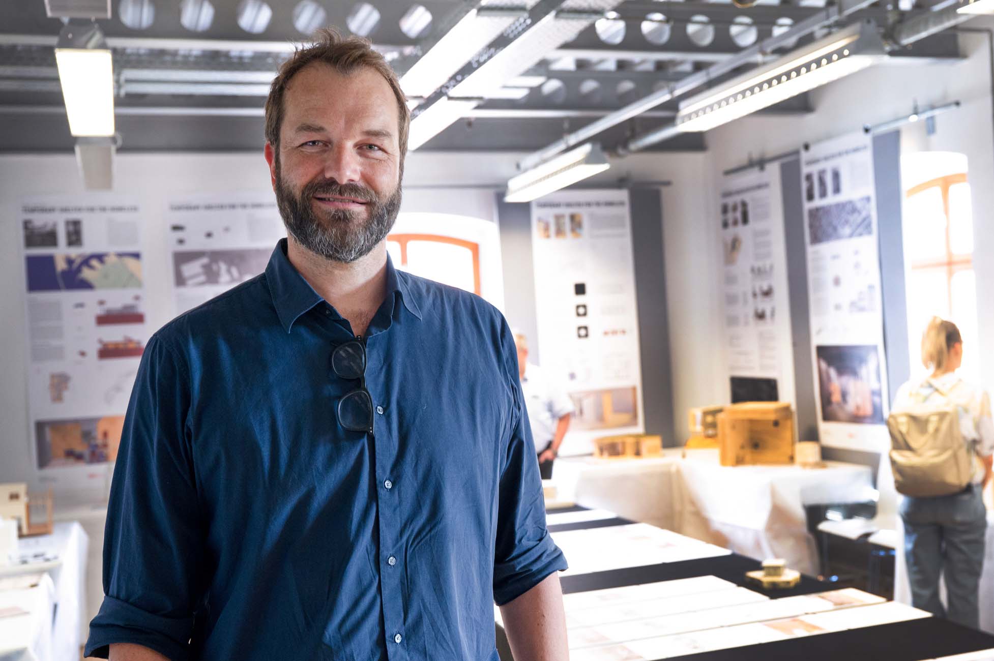 Ein Mann mit Bart und blauem Hemd steht lächelnd in einem hellen Raum mit Ausstellungsplakaten und Tischen, auf denen verschiedene Gegenstände ausgestellt sind. Im Hintergrund schauen sich andere Leute die Exponate an. at Hochschule Coburg