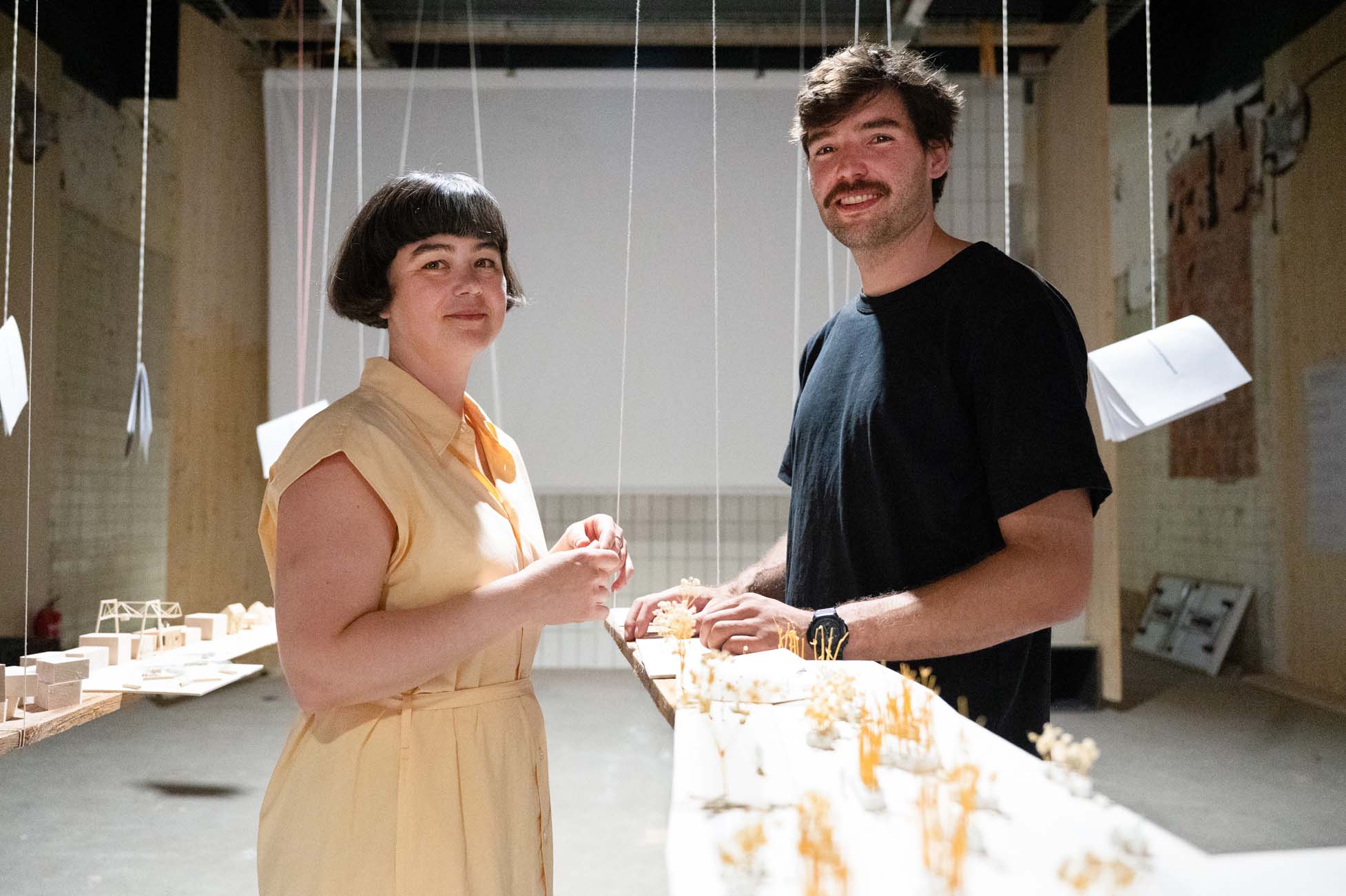 Eine Frau und ein Mann stehen in einem hellen Raum mit hängenden Schnüren und Architekturmodellen auf einem Tisch vor ihnen. Beide lächeln und schauen in die Kamera. Der Raum hat eine kreative, studioähnliche Atmosphäre. at Hochschule Coburg