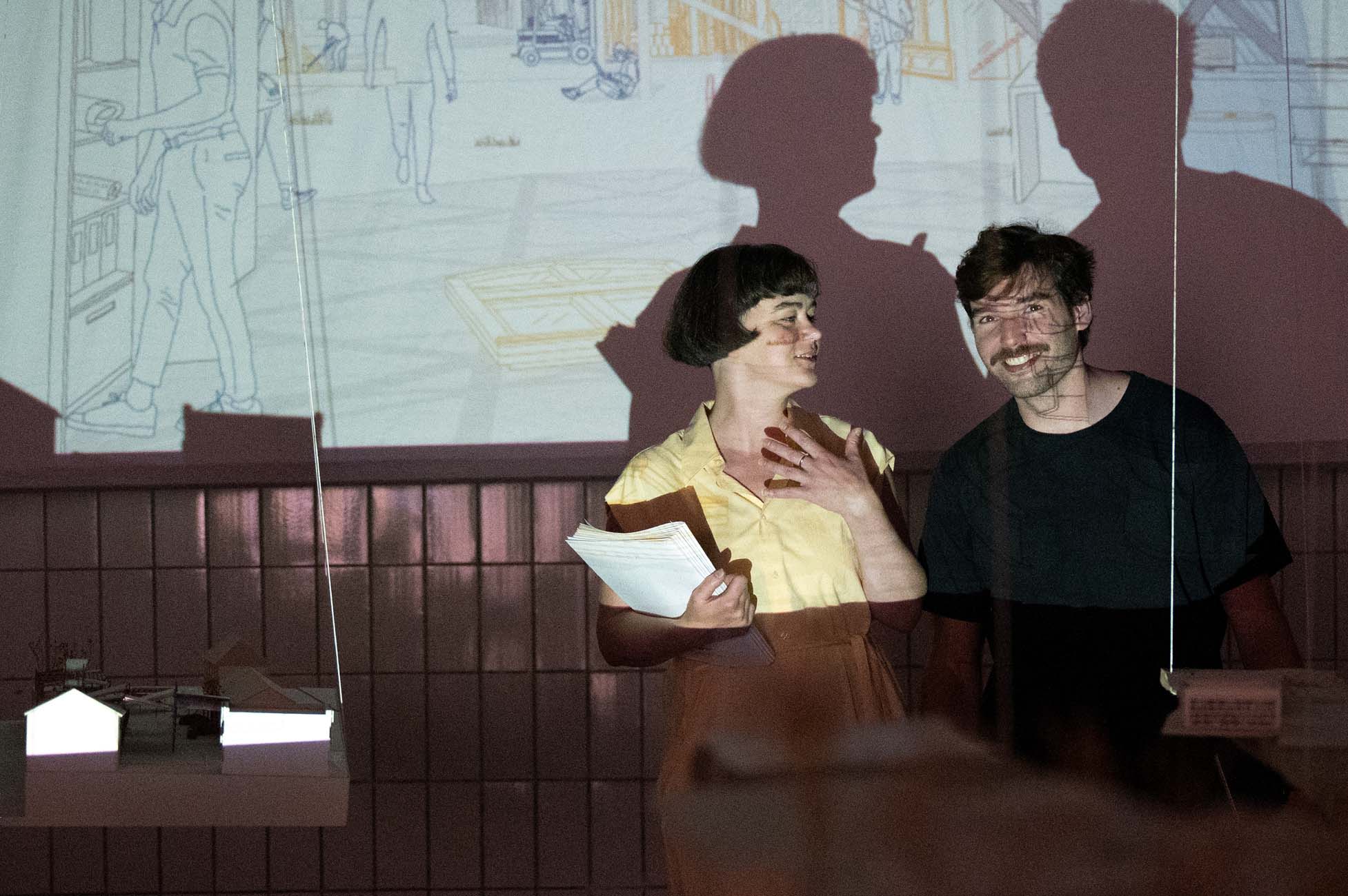 Zwei Personen stehen lächelnd vor einer Wand mit projizierten Zeichnungen. Einer hält Papiere in der Hand, und beide haben ihre Schatten hinter sich. Auf einem Tisch im Vordergrund befinden sich kleine Modelle. at Hochschule Coburg