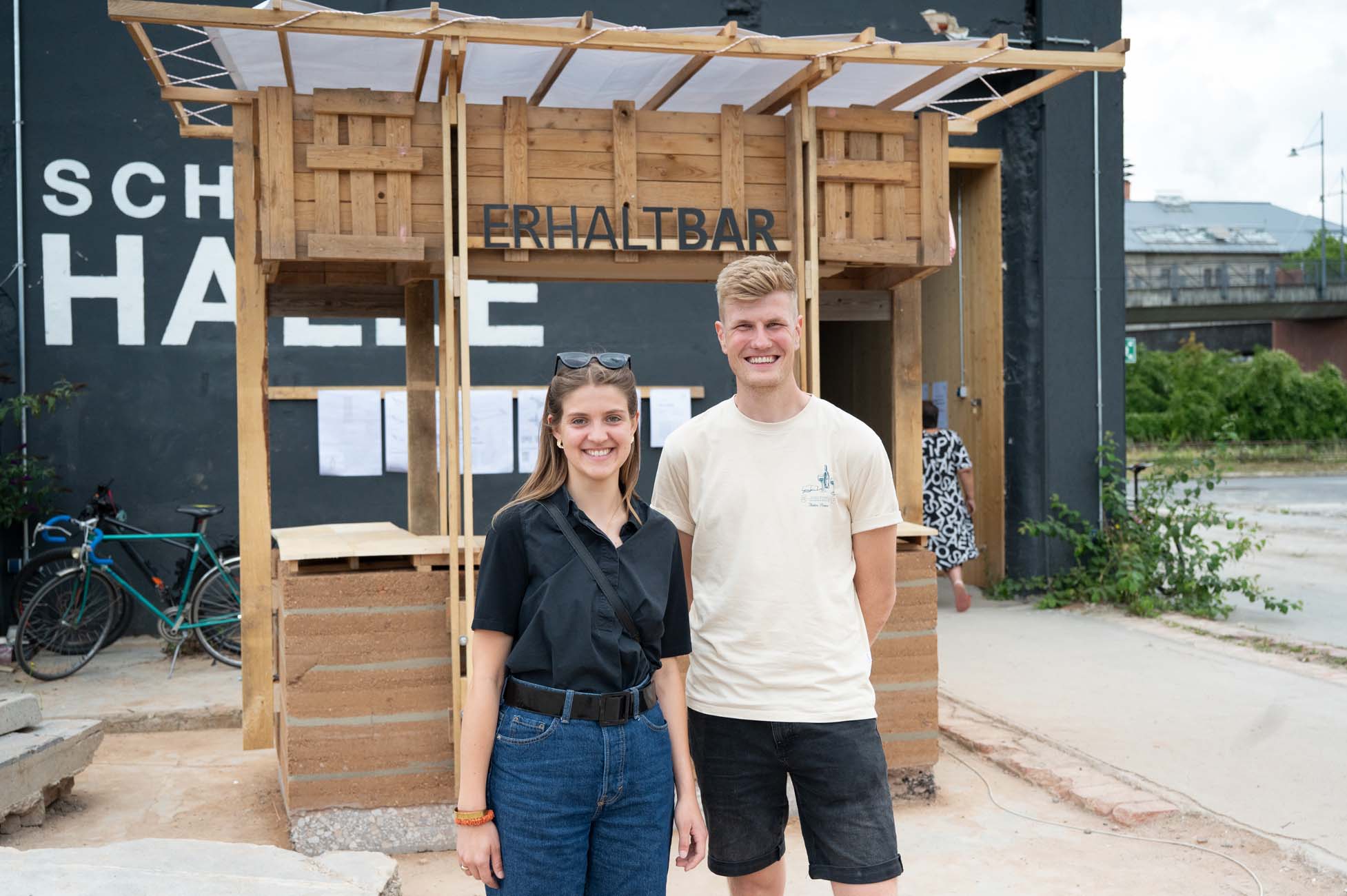 Zwei junge Erwachsene stehen lächelnd vor einer kleinen Holzkonstruktion mit der Aufschrift "ERHALTBAR". Im Hintergrund sind ein schwarzes Gebäude, ein Fahrrad und etwas Grün zu sehen. Beide Personen sind leger gekleidet. at Hochschule Coburg