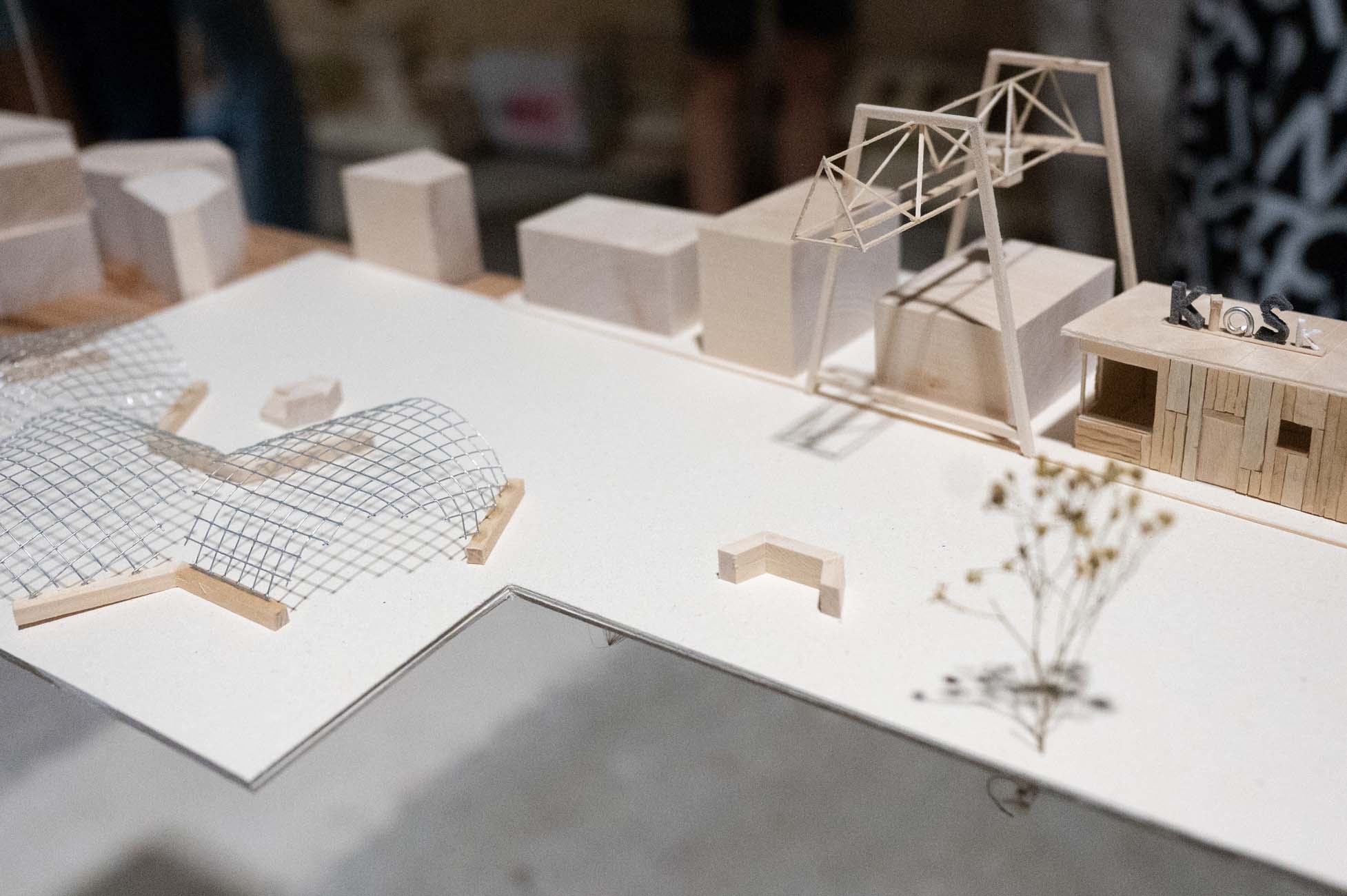 Nahaufnahme eines Architekturmodells mit kleinen Holzstrukturen, einer Kuppel aus Drahtgitter, einem Kiosk und abstrakten kastenförmigen Gebäuden auf weißem Grund. In der Nähe der rechten Seite des Modells befindet sich eine getrocknete Pflanze. at Hochschule Coburg