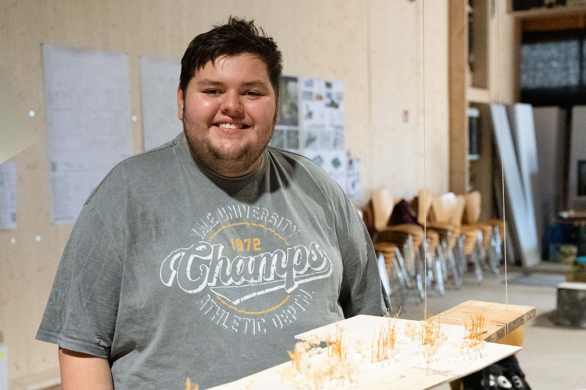 Eine lächelnde Person in einem grauen "Champs"-T-Shirt steht in einem Raum und hält ein Architekturmodell in der Hand. Dahinter befinden sich gestapelte Stühle, ein schwarzes Brett und an die Wand gepinnte Papiere. at Hochschule Coburg